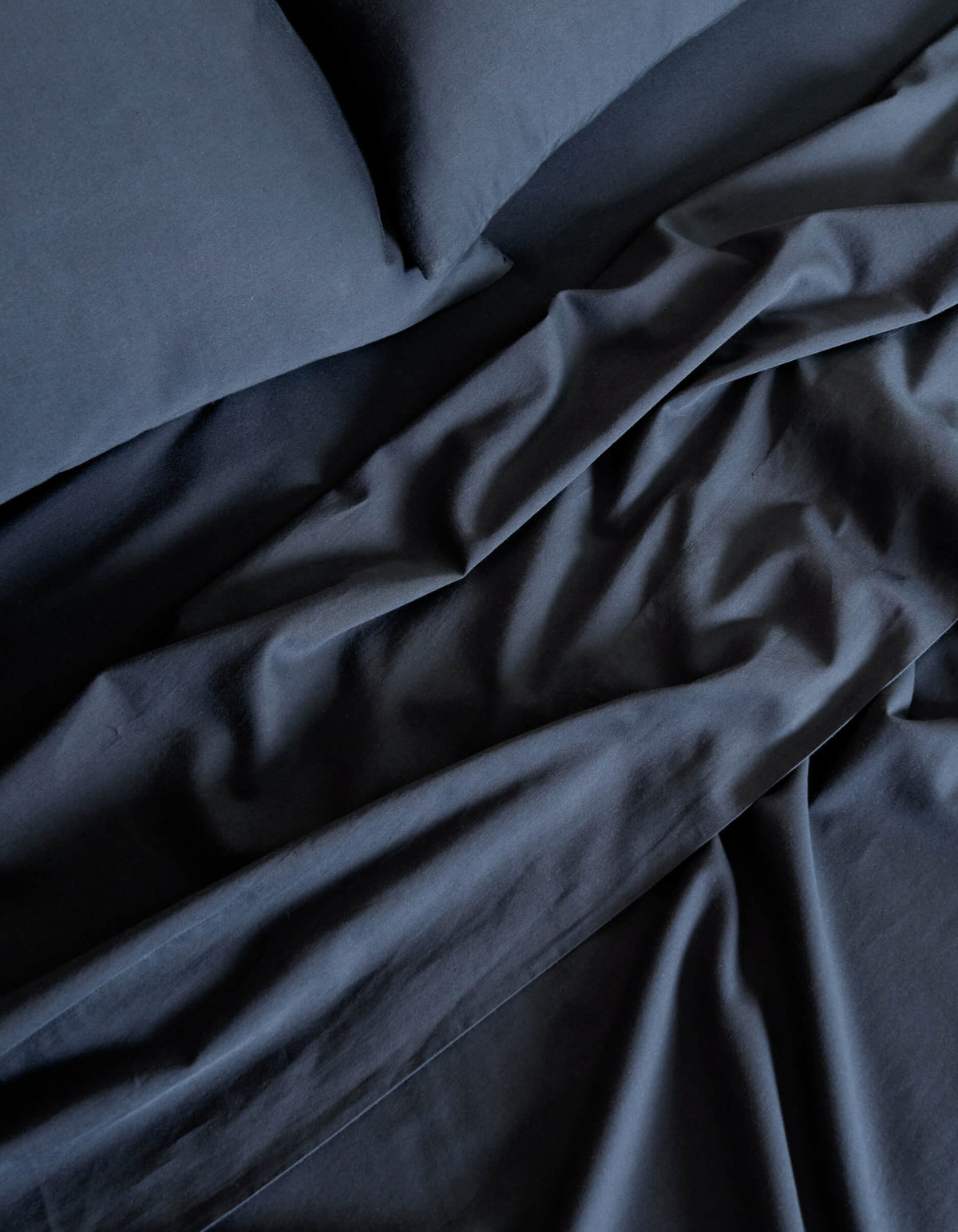 A close-up of the Cozy Earth Soft-Wash Cotton Sheet Set in dark blue highlights the soft folds and creases in the fabric on the bed.|Color:Eclipse