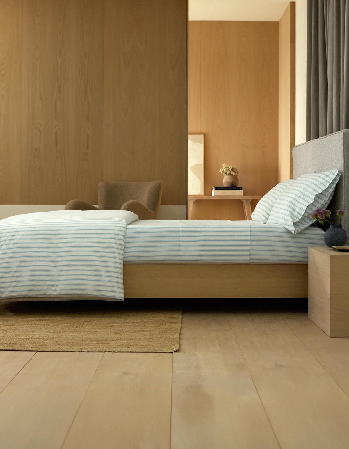A modern, minimalist bedroom featuring a low wooden bed with Cozy Earth’s Soft-Wash Cotton Fitted Sheet, striped bedding, a beige rug, gray headboard, wooden nightstand with small vases, armchair, and light wood paneling.|Color:River and Sea Salt Stripe