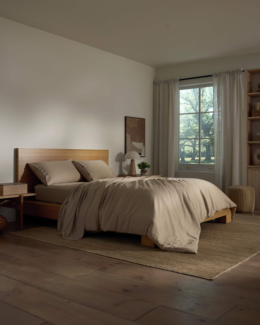 A cozy bedroom featuring a wooden bed frame with Cozy Earth's Soft-Wash Cotton Bedding Deluxe Bundle in beige, a nightstand with a lamp and plant, sheer curtains letting in natural light, and wooden floors topped by a woven rug.|Color:Sandalwood