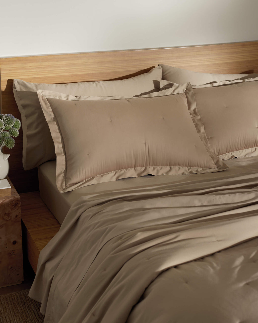 A neatly made bed features Cozy Earth's Soft-Wash Cotton Quilted Shams in beige, paired with matching sheets and a duvet against a wooden headboard. A small nightstand with a decorative plant completes the calm and cozy ambiance.|Color:Sandalwood