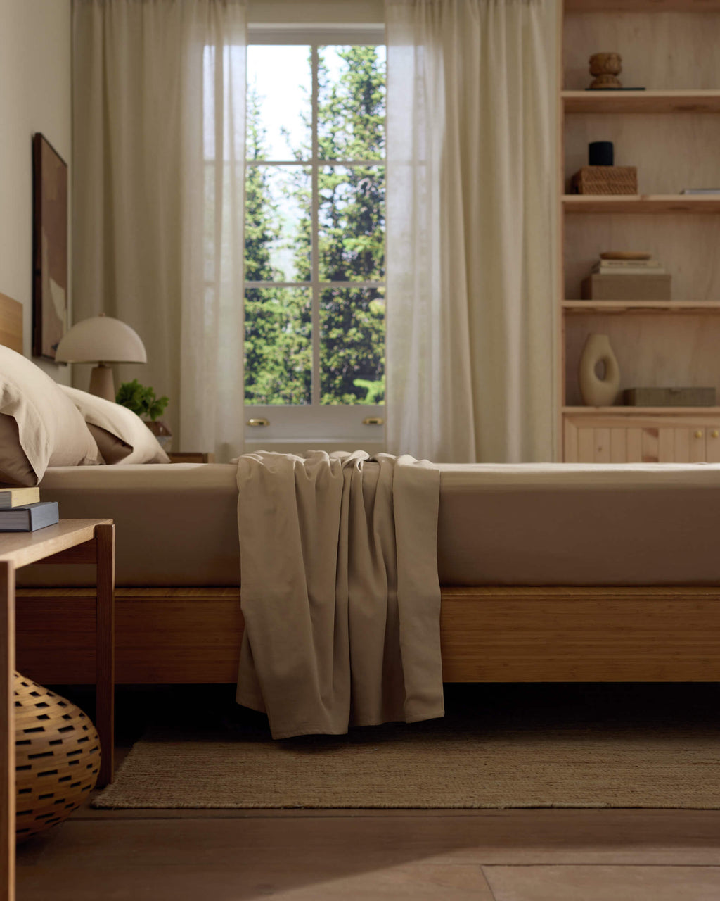 A sunlit bedroom features a neatly made bed with Cozy Earth's Soft-Wash Cotton Sheet Set, beige bedspread, wooden furniture, decor shelves, and a large window with sheer curtains framing green trees outside.|Color:Sandalwood