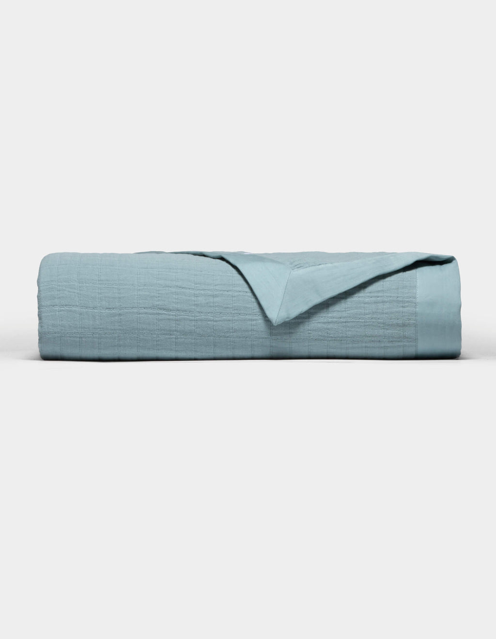 The Cozy Earth Waterfront Grid Matelasse Blanket in light blue is neatly folded against a plain white background, with the top layer turned back to show the inner fabric.|Color:River
