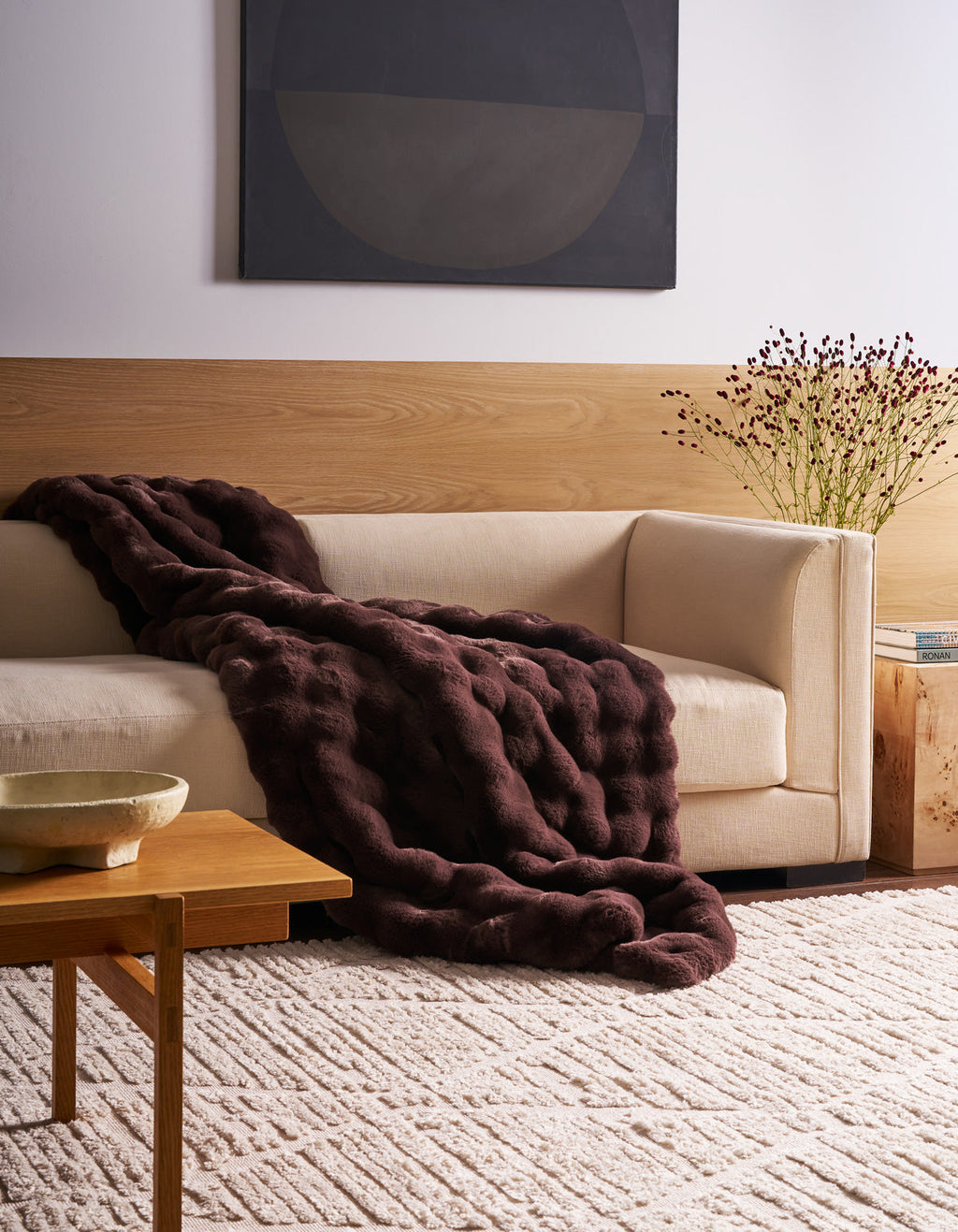 A modern living room featuring a cream-colored sofa, adorned with the Cozy Earth Bubble Cuddle Blanket, complemented by a dark purple textured blanket, a wooden table, a patterned rug, and minimalist decor.|Color:Raisin