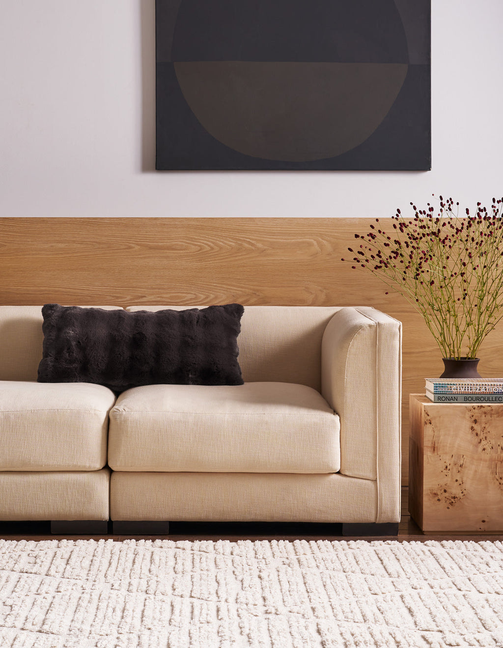 A beige sofa with a Cozy Earth Bubble Cuddle Pillow sits beside a wooden side table holding books and a vase with twigs. A textured white rug covers the floor, and abstract art hangs on the wall above wood paneling.|Color:Raisin |Size:14" x 30"