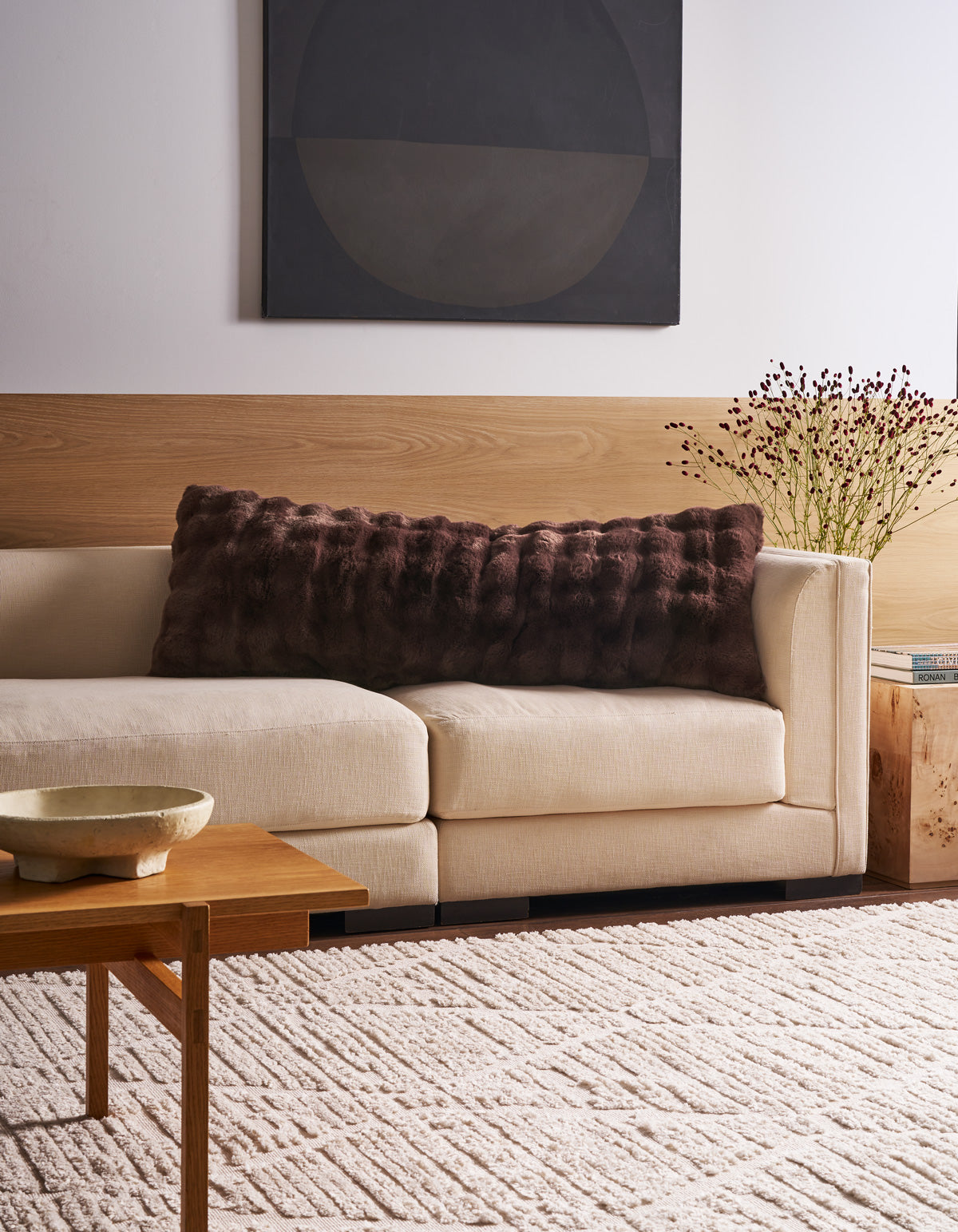 A modern living room featuring a cream sofa adorned with the Bubble Cuddle Pillow by Cozy Earth, abstract wall art, a wooden coffee table with a decorative bowl, a side table topped with books and dried flowers, and a patterned beige rug.|Color:Raisin |Size:Body - 20" x 54"