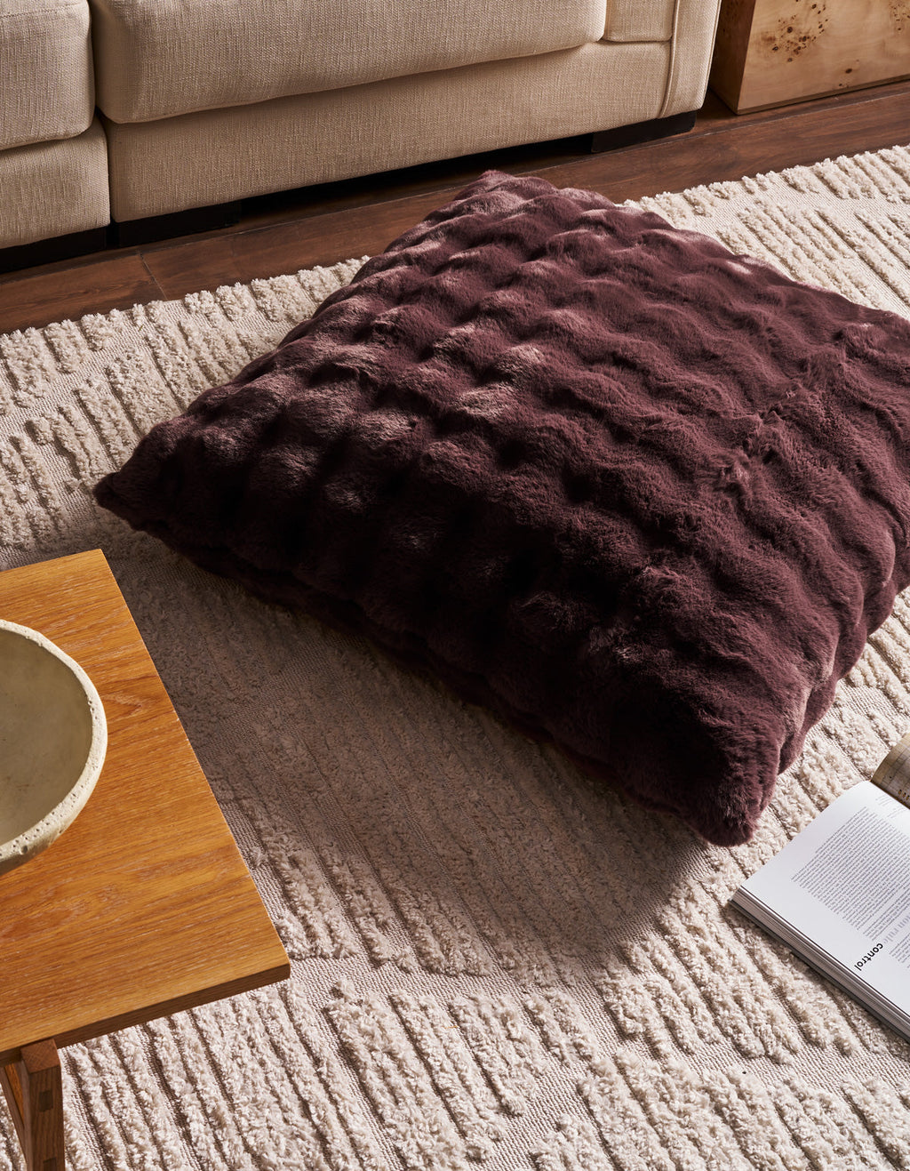 A spacious, textured Bubble Cuddle Pillow by Cozy Earth in dark brown rests atop a cream patterned rug beside a wooden coffee table and an open magazine, creating a cozy living room atmosphere.|Color:Raisin |Size:36" x 36"