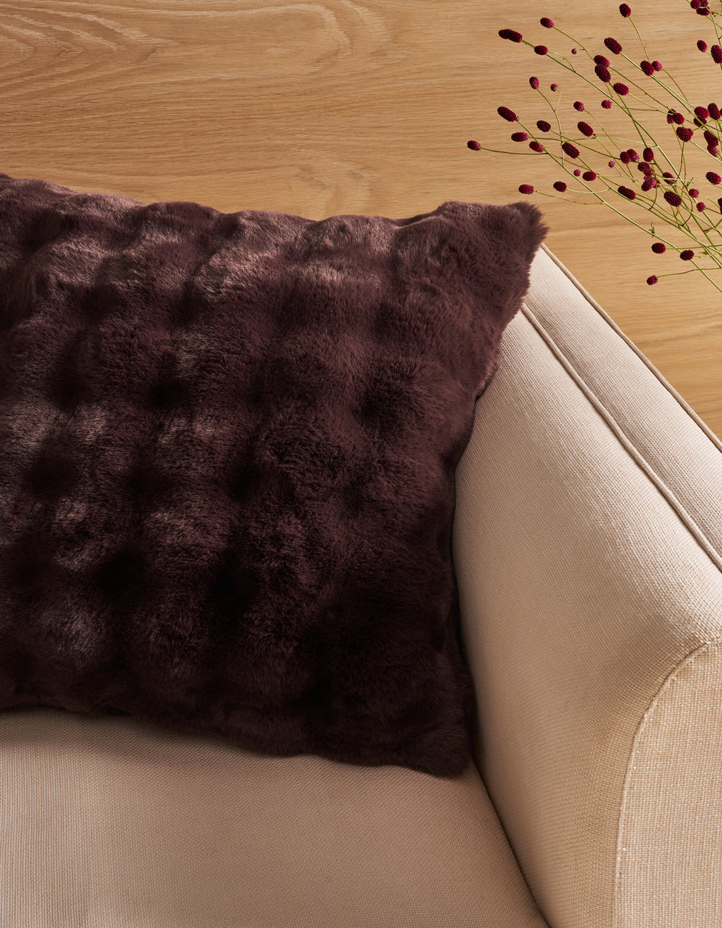 A Bubble Cuddle Pillow by Cozy Earth, featuring a dark brown textured faux fur design, rests on a beige armchair beside a light wooden surface with delicate red berry stems in the background.|Color:Raisin |Size:26" x 26"