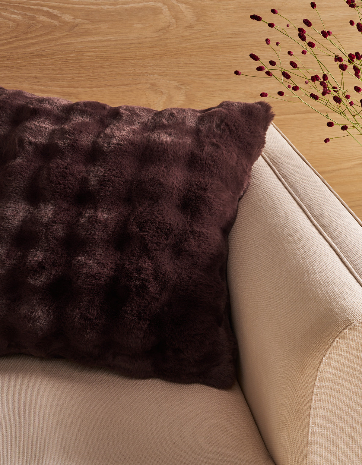 A Bubble Cuddle Pillow by Cozy Earth, featuring a dark brown textured faux fur design, rests on a beige armchair beside a light wooden surface with delicate red berry stems in the background.|Color:Raisin |Size:26" x 26"