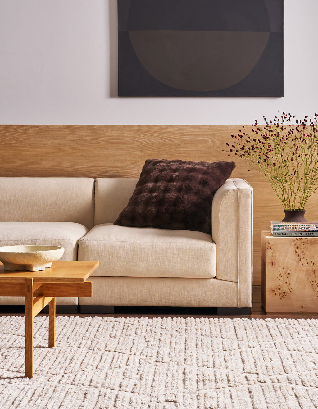 A modern living room with a beige sofa, a Cozy Earth Bubble Cuddle Pillow in a dark textured finish, a wooden coffee table, light rug, side table with books and a vase of flowers, and abstract art on the wall.|Color:Raisin
