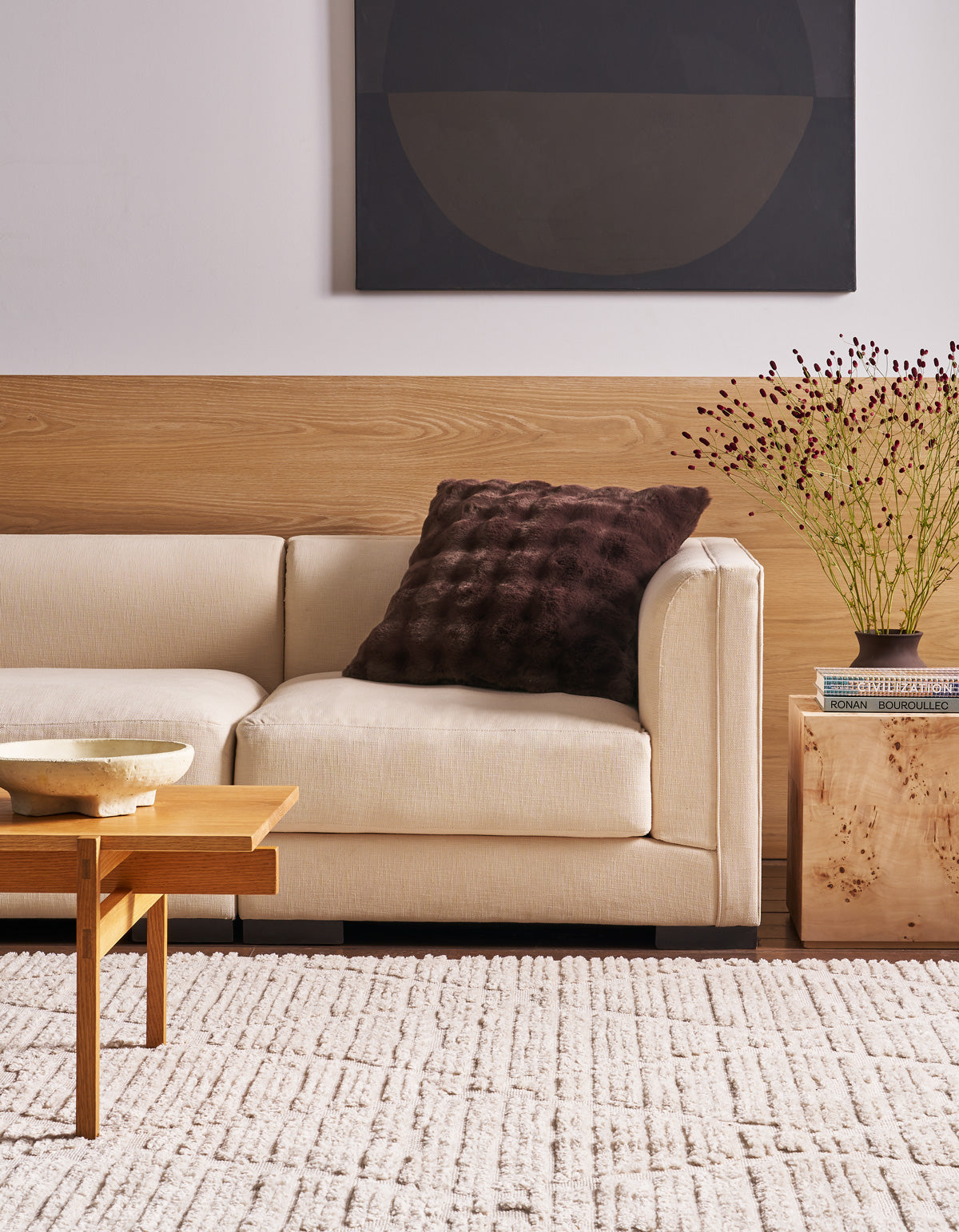 A modern living room with a beige sofa, a Cozy Earth Bubble Cuddle Pillow in a dark textured finish, a wooden coffee table, light rug, side table with books and a vase of flowers, and abstract art on the wall.|Color:Raisin
