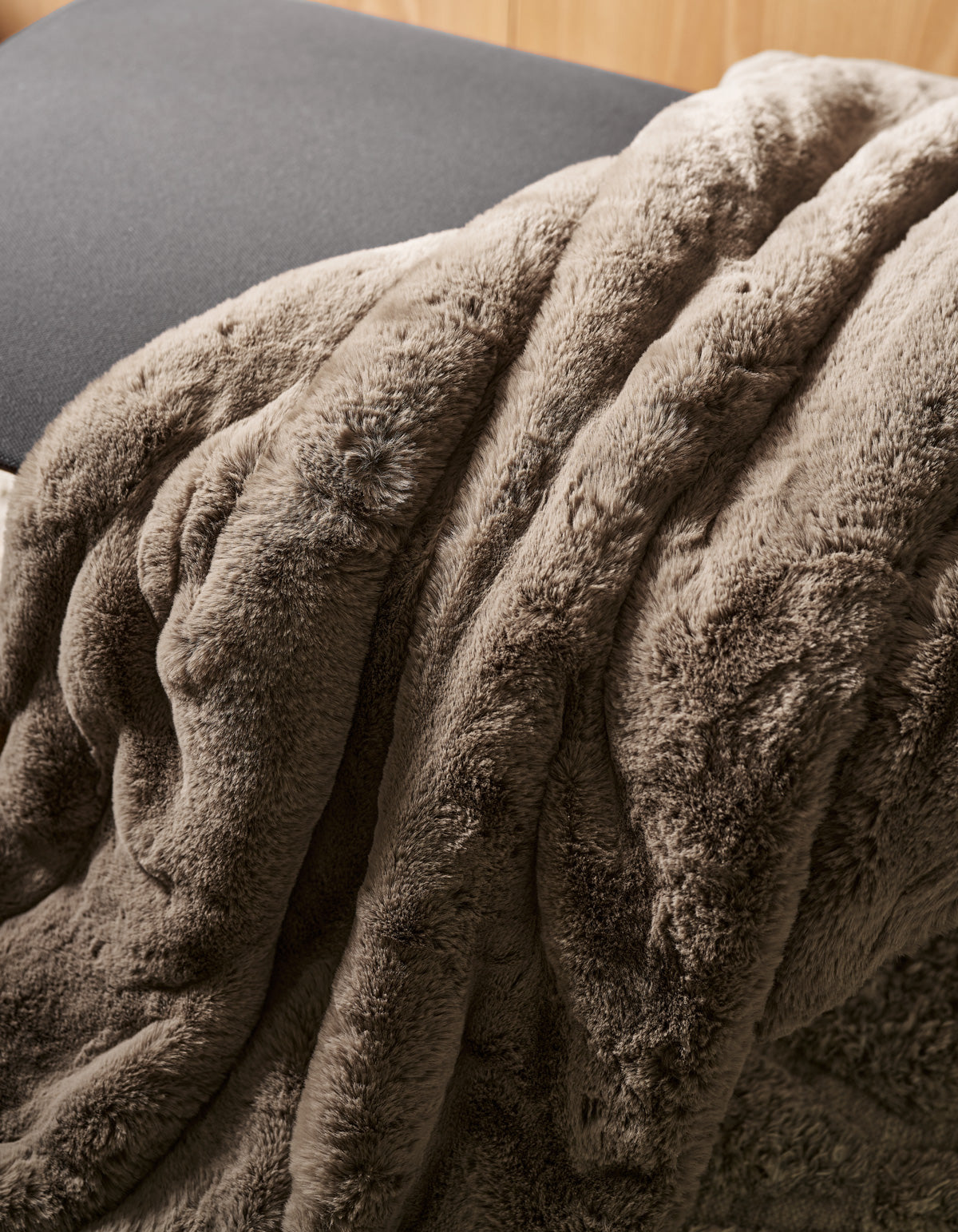 The Channel Cuddle Blanket by Cozy Earth, in a soft brown plush fabric, is draped over the edge of a gray cushion or chair, creating a cozy and inviting atmosphere.|Color:Walnut