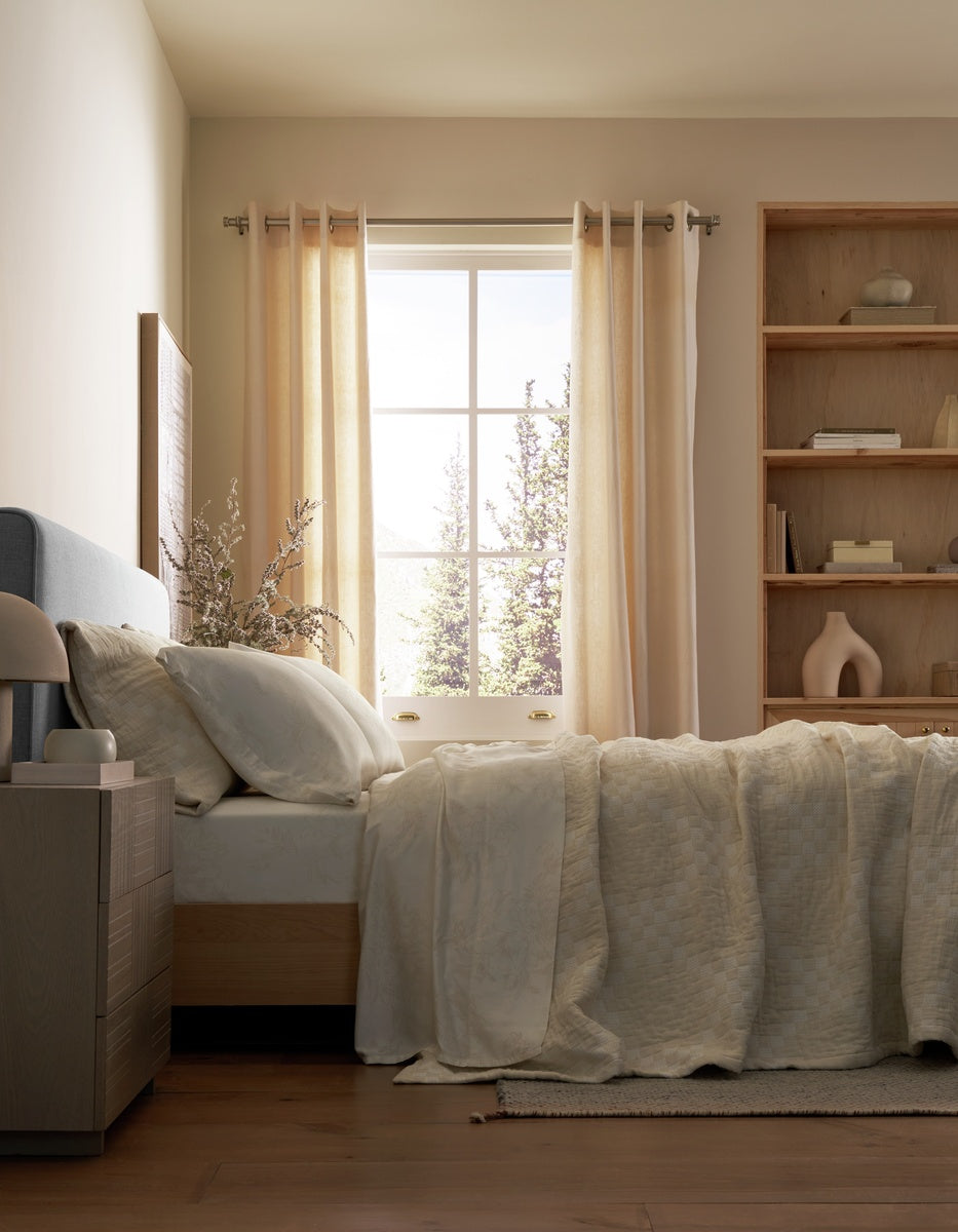 A neatly made bed with the Cozy Earth Sonoma Stitch Quilt in cream is positioned beside a window dressed in sheer curtains. Sunlight fills the room, highlighting a wooden nightstand and built-in shelving adorned with decorative objects.|Color:Vintage White