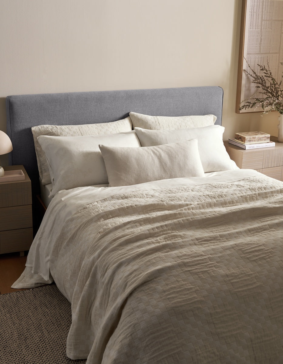 A neatly made bed features a gray upholstered headboard, cream-colored bedding, multiple pillows including the Sonoma Throw Pillow by Cozy Earth, and a beige quilt. Beside the bed, a nightstand holds a lamp and books, while decorative accents adorn the walls and tabletop.|Color:Canvas|Size:14" x 30"
