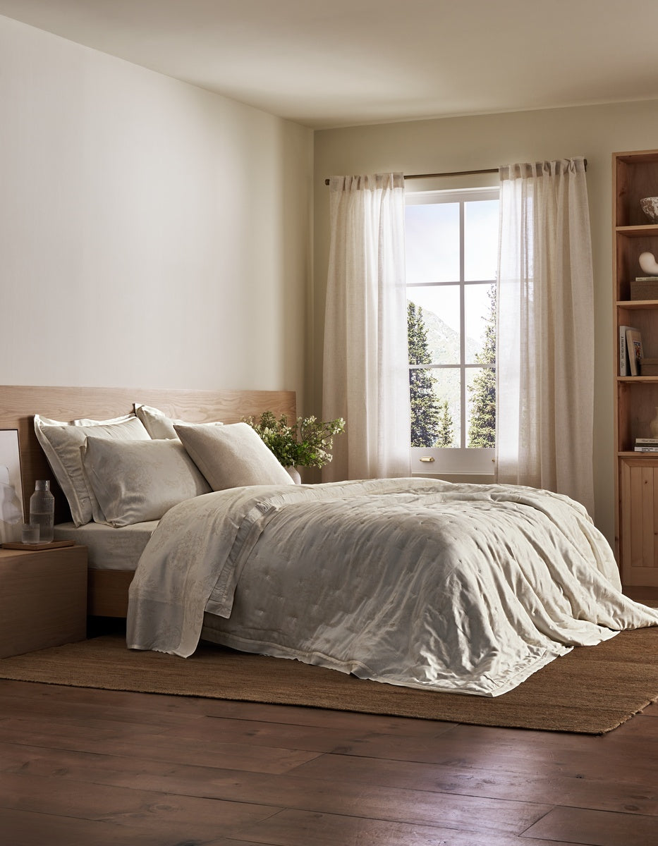 A cozy bedroom features a neatly made bed adorned with the Sonoma Throw Pillow from Cozy Earth, light bedding, a wooden nightstand, and a bookcase. Sunlight streams through sheer curtains on the window, illuminating the room’s neutral tones and hardwood floor.|Color:Canvas|Size:24" x 24"