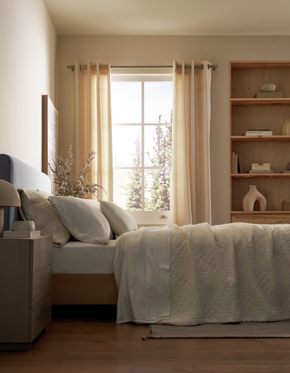 A sunlit bedroom features a neatly made bed adorned with light beige bedding and a Cozy Earth Sonoma Throw Pillow. A wooden nightstand sits nearby, while shelves decorated with tasteful accents and large windows with sheer curtains let in abundant natural light.|Color:Sea Salt|Size:14" x 30"