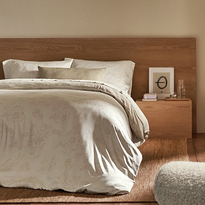 A cozy bedroom featuring a wooden bed with light bedding and accented by the Cozy Earth Sonoma Throw Pillow. A nightstand holds books and a glass of water, while sunlight streams through beige curtains on a large window. Wooden floors and shelves enhance the room’s warmth.|Color:Tallow|Size:14" x 30"