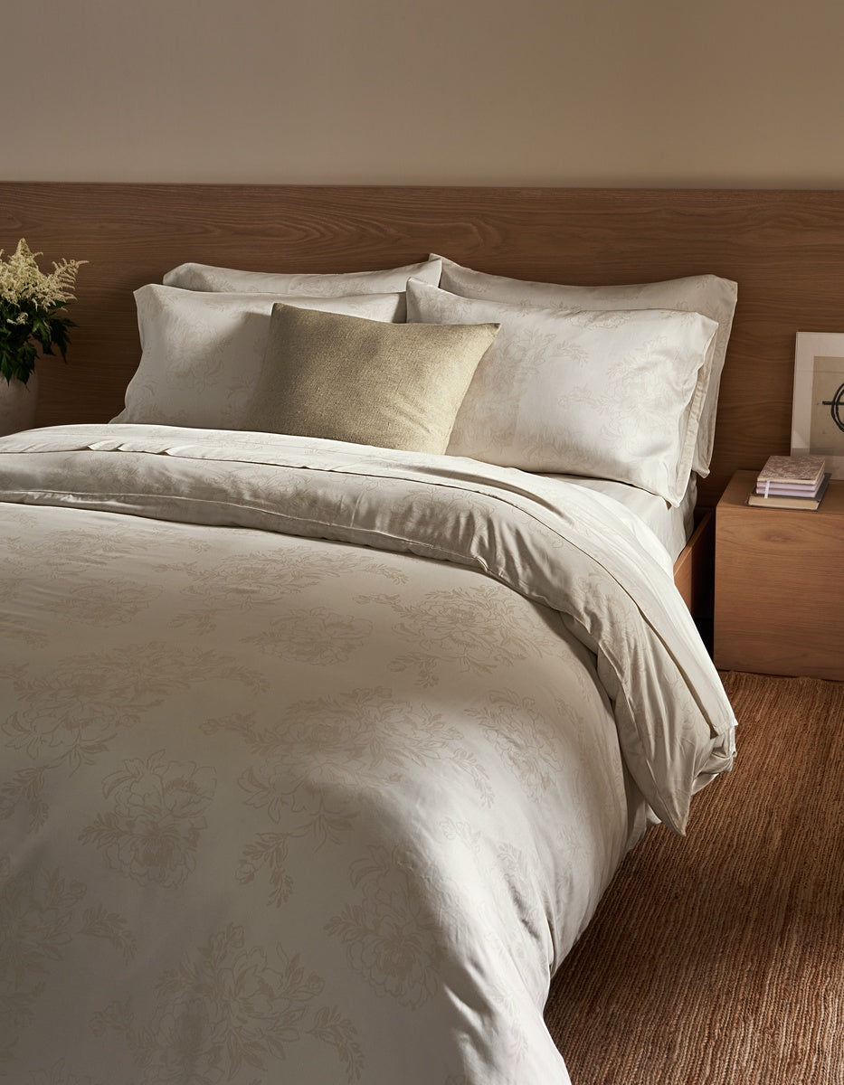 A neatly made bed with cream-colored bedding and a single Cozy Earth Sonoma Throw Pillow in beige, next to a wooden nightstand with stacked books and a small flower arrangement.|Color:Tallow|Size:24" x 24"