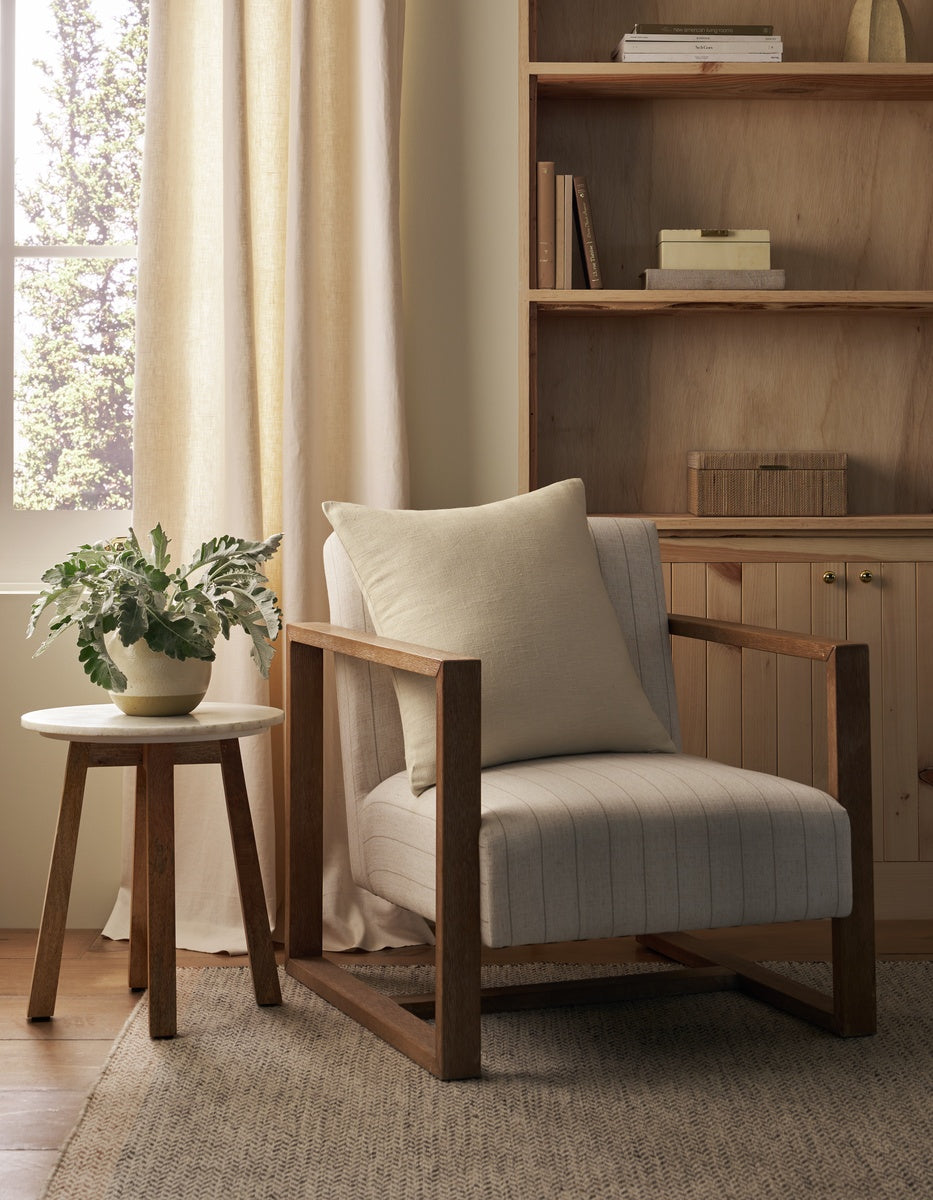 A cozy reading nook with a wooden armchair, a Cozy Earth Sonoma Throw Pillow, a round side table topped with a potted plant, and built-in wooden shelves in a softly lit room.|Color:Canvas|Size:26" x 26"