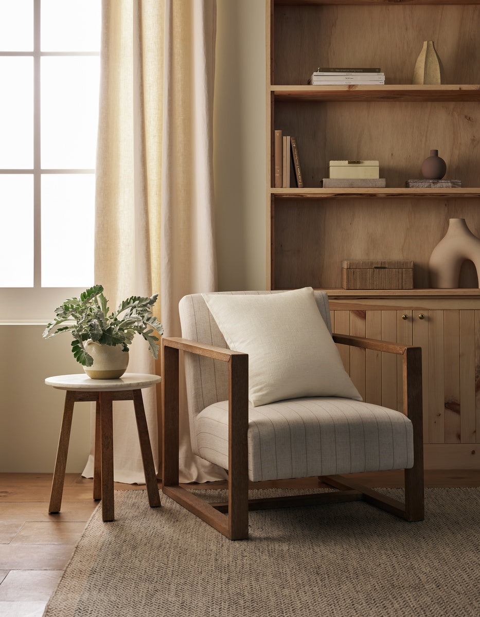 A cozy reading nook featuring a wooden armchair topped with the Cozy Earth Sonoma Throw Pillow, a round side table holding a potted plant, built-in shelves, and a window adorned with light curtains that let in soft natural light.|Color:Sea Salt|Size:26" x 26"