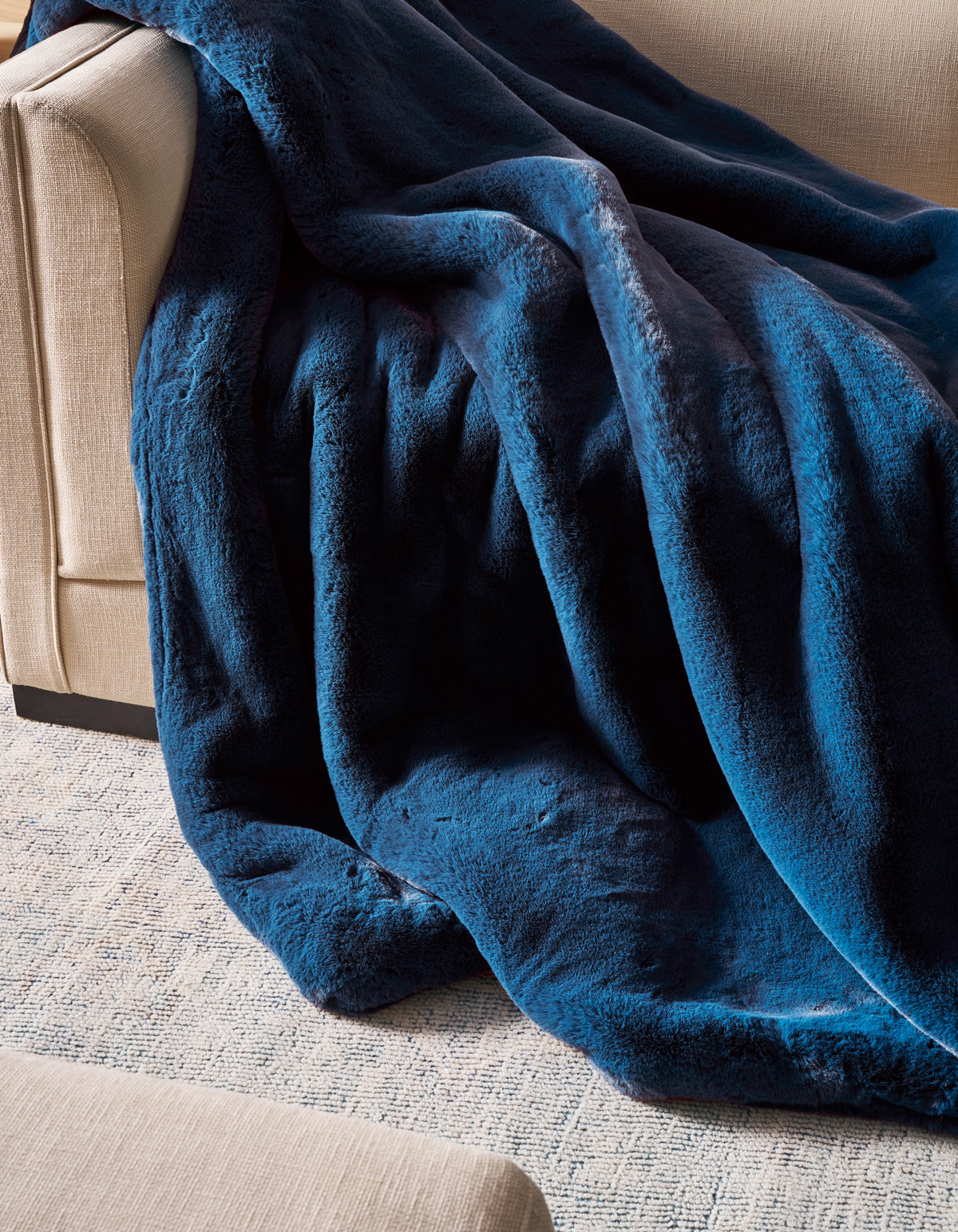 A Superplush Cuddle Blanket by Cozy Earth in rich dark blue is draped over the arm and seat of a beige sofa, with part of the blanket pooling onto a light-colored textured rug.|Color:Deep Water