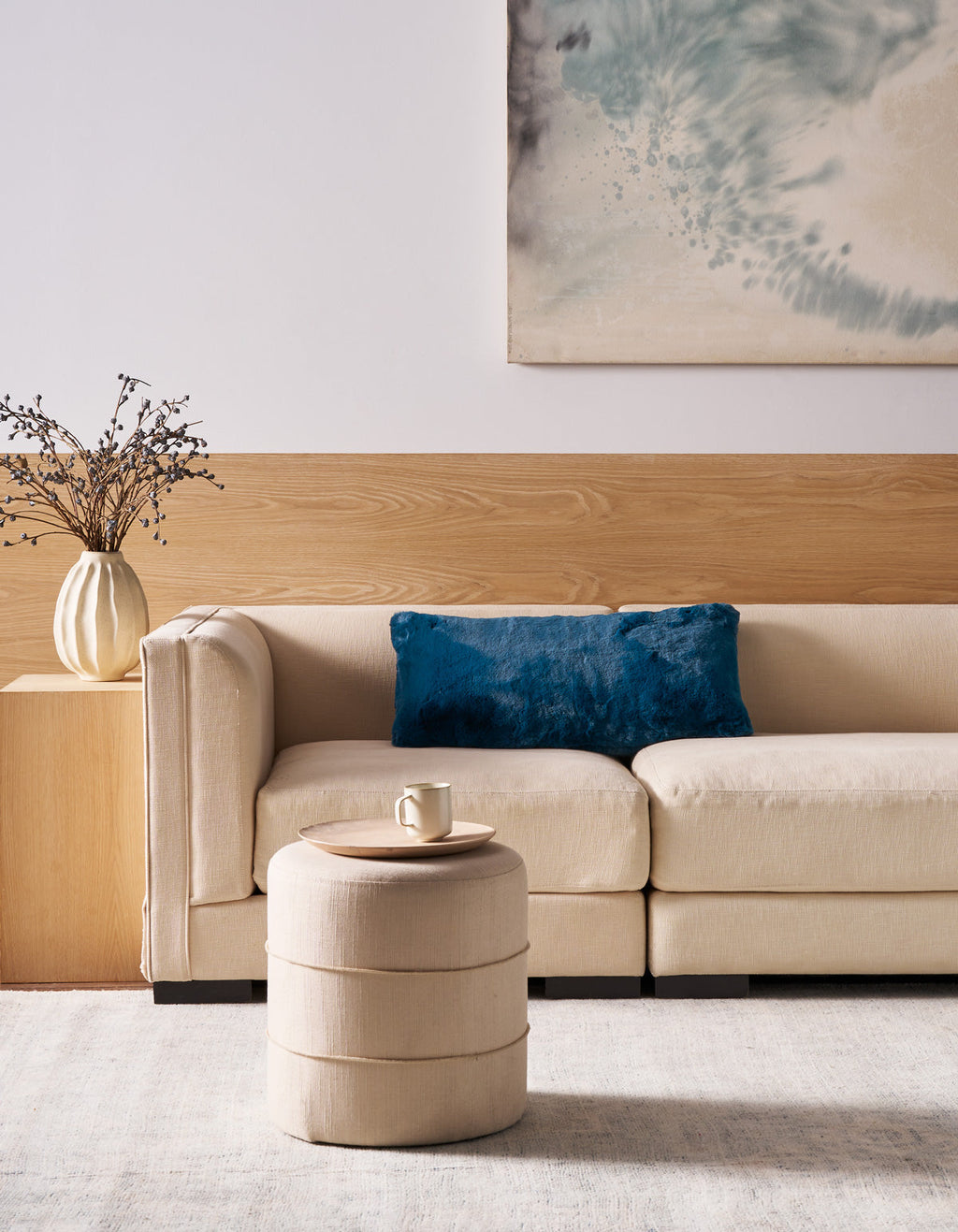 A modern living room with a beige sofa, a Superplush Cuddle Pillow by Cozy Earth in blue, a round beige ottoman holding a mug, a white vase with branches, and abstract art on a wood and white wall.|Color:Deep Water |Size:14" x 30"