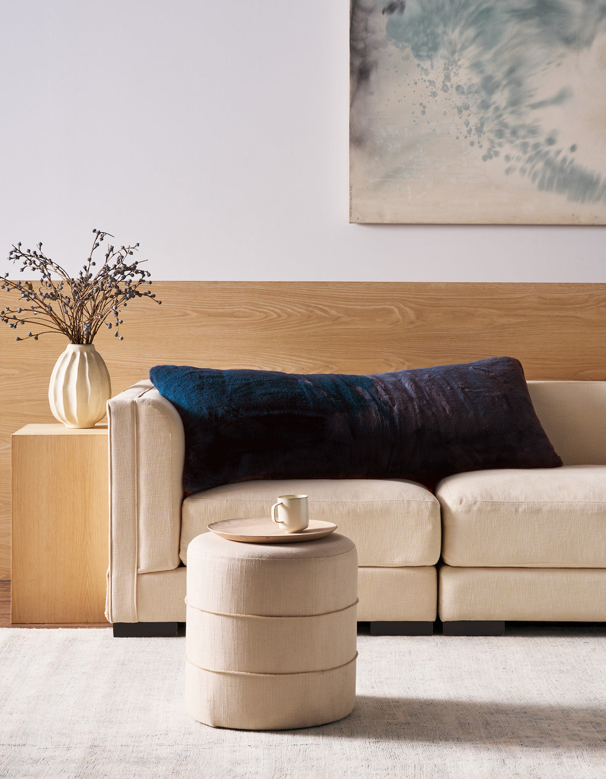 A modern living room features a beige sofa with the Superplush Cuddle Pillow by Cozy Earth in large dark blue, a round beige ottoman with a mug, a wooden side table with a vase of branches, and abstract artwork on the wall.|Color:Deep Water |Size:Body - 20" x 54"