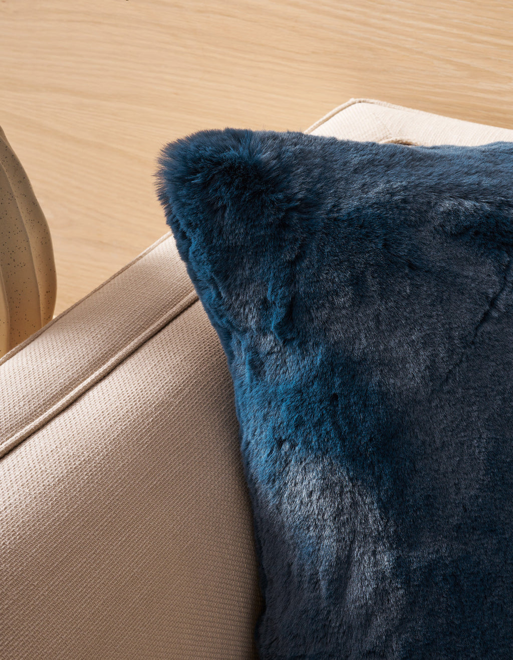 A close-up of a beige fabric couch featuring the Superplush Cuddle Pillow by Cozy Earth in dark blue faux fur, resting on the corner and set against a light wooden background.|Color:Deep Water |Size:26" x 26"