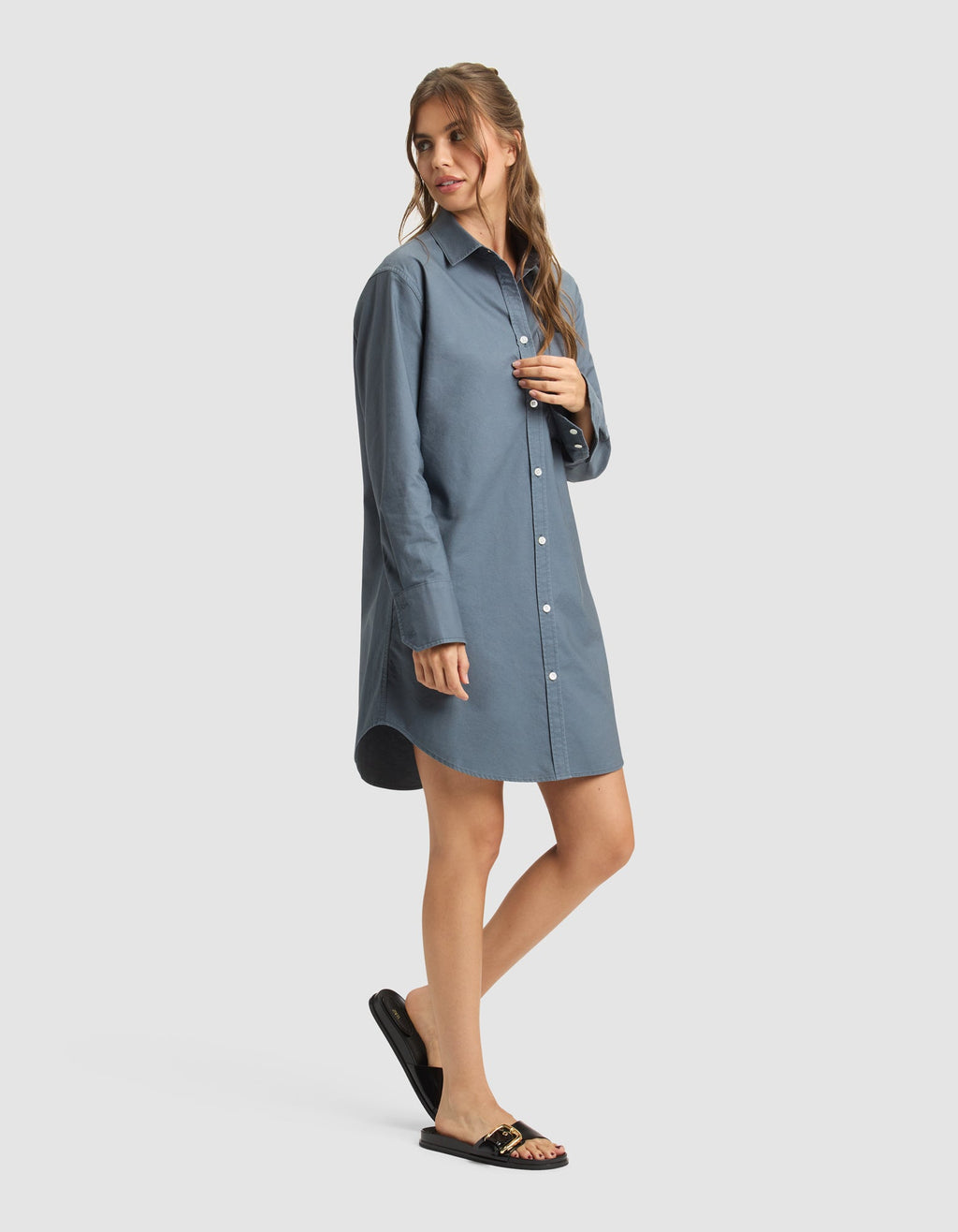 A woman poses in a relaxed way, wearing Cozy Earth's Women's Camden Dress—a loose-fitting, long-sleeve blue shirt dress—paired with black sandals featuring gold accents against a plain light background. |Color:Dawn