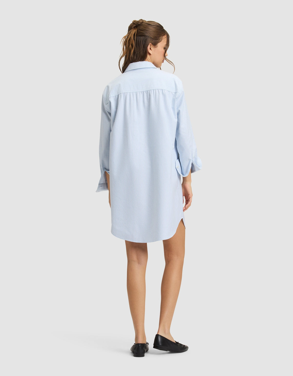 A woman with long brown hair in a ponytail stands facing away, wearing the Cozy Earth Women's Camden Dress—a light blue, loose-fitting, long-sleeved shirt dress—with black shoes against a plain light gray background. |Color:Blue Oxford Stripe