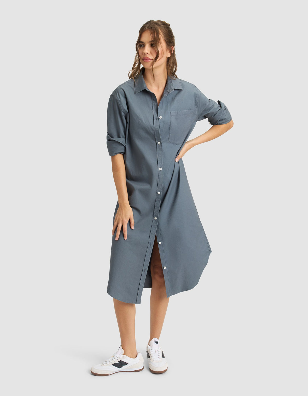 A woman stands against a light background wearing the Cozy Earth Women's Camden Midi Dress, a loose blue button-down style with rolled-up sleeves, paired with white sneakers. Her light brown hair is tied back as she poses with one hand on her hip. |Color:Dawn