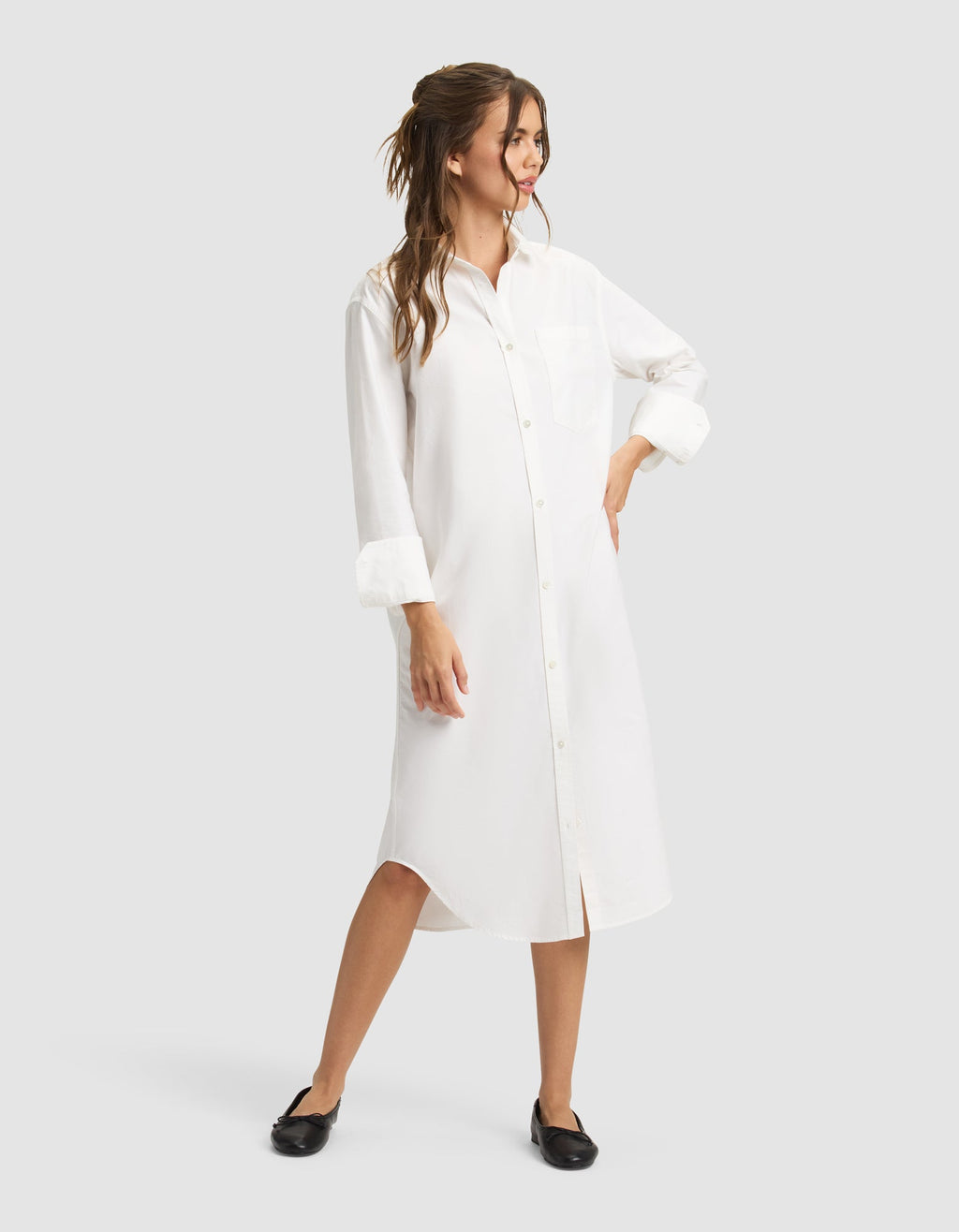 A woman wearing the Cozy Earth Women's Camden Midi Dress with rolled-up sleeves and black flats smiles, looking to the side with wavy brown hair styled half-up and one hand on her hip. |Color:White