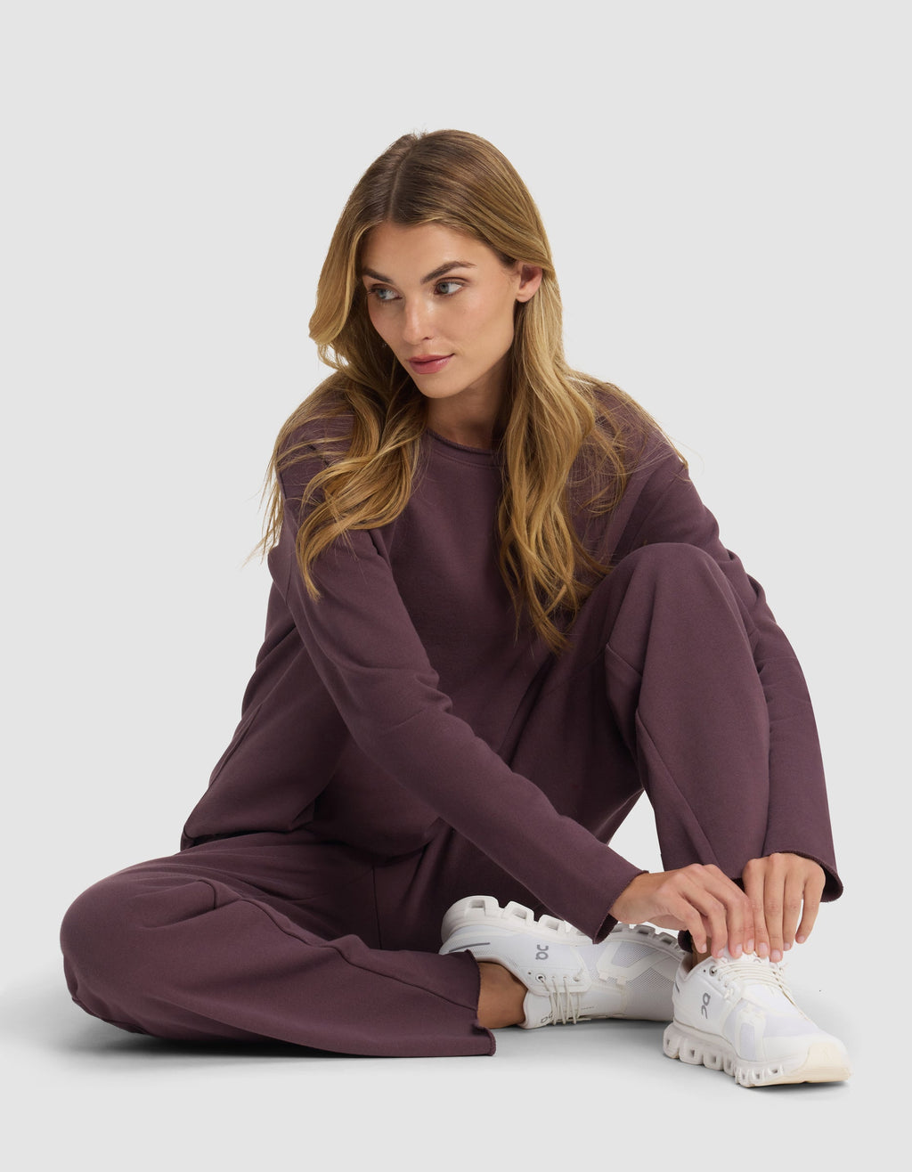 A woman with long blonde hair sits on the floor wearing a loose-fitting mauve Chelsea Pant by Cozy Earth and white sneakers, looking to her right while adjusting her shoe against a plain light background.|Color:Fig