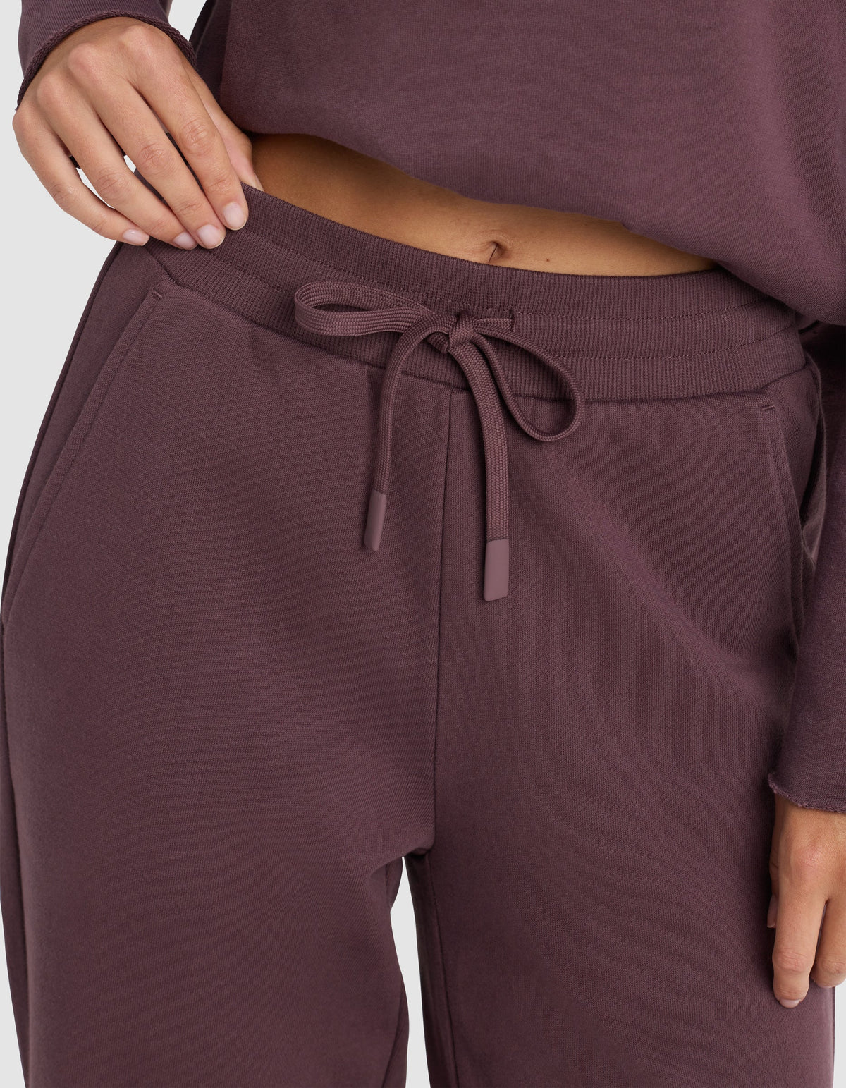 A person wears Cozy Earth's Chelsea Pant, a soft and comfortable drawstring sweatpant, in matching mauve or dark purple, with their hand resting on the waistband.|Color:Fig