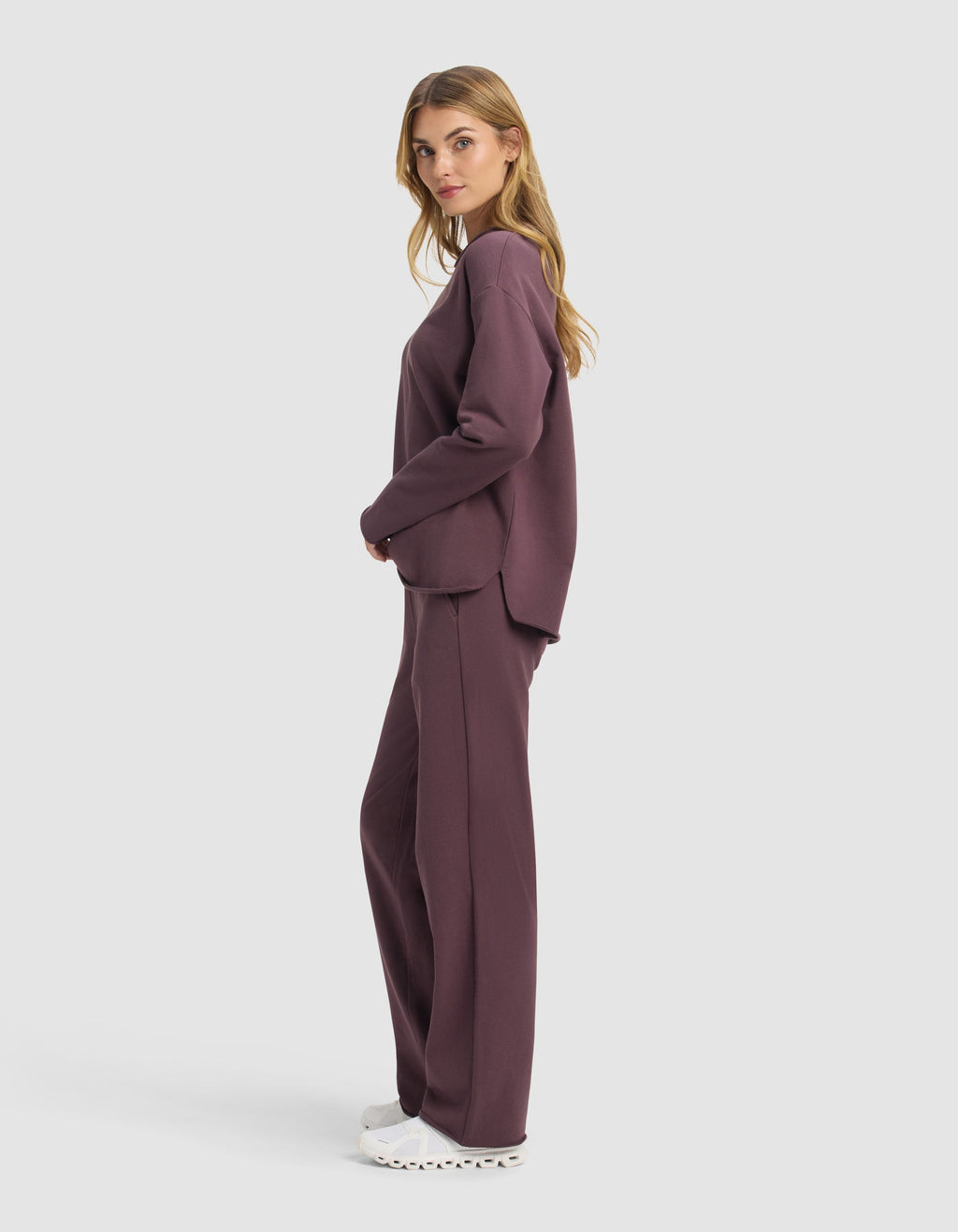 A woman stands in profile wearing the Cozy Earth Women's Chelsea Set—loose-fitting, long-sleeve dark mauve sweatshirt and matching pants—with white sneakers, set against a plain light background.|Color:Fig