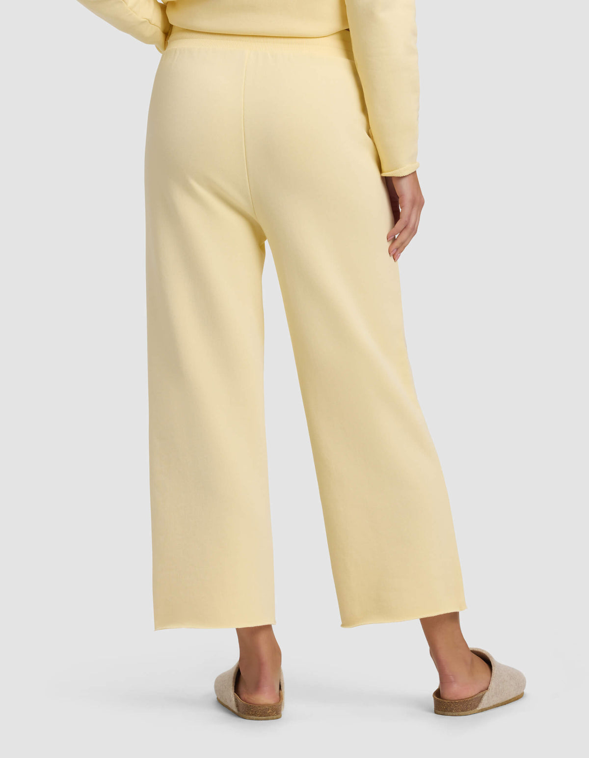 A person is seen from the back wearing Cozy Earth's Women's Chelsea Cropped Pant in light yellow with a matching top, standing on a white surface and beige slip-on slippers.|Color:Meringue
