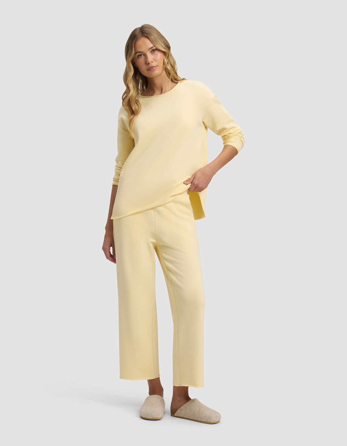 A woman with long blonde hair wears Cozy Earth's Women's Chelsea Cropped Pant in pale yellow with a matching loose top and beige slippers, standing against a plain light gray background.|Color:Meringue