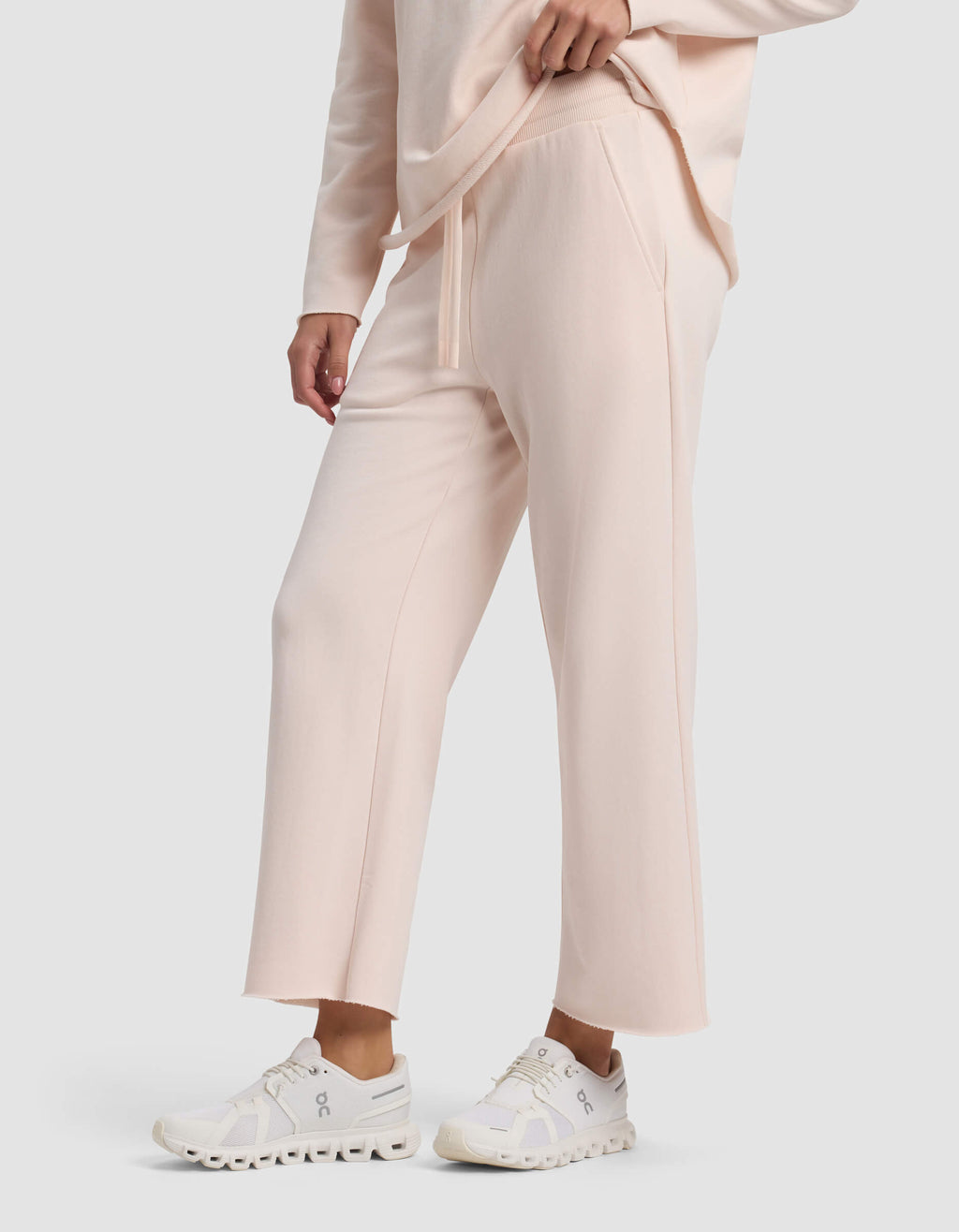 A person wears Cozy Earth's Women's Chelsea Cropped Pant in light pink with a matching sweatshirt and white athletic sneakers, standing with one hand in their pocket against a plain light gray background.|Color:Petal