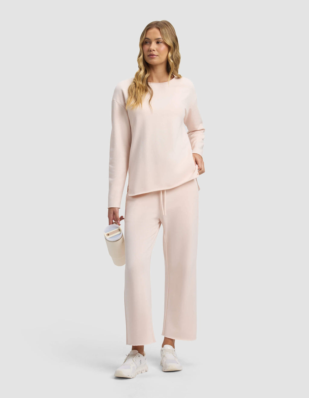 A woman with long wavy blonde hair wears Cozy Earth's Women's Chelsea Cropped Pant and a light pink long-sleeve top, paired with white sneakers, holding a white tumbler against a plain light gray background.|Color:Petal