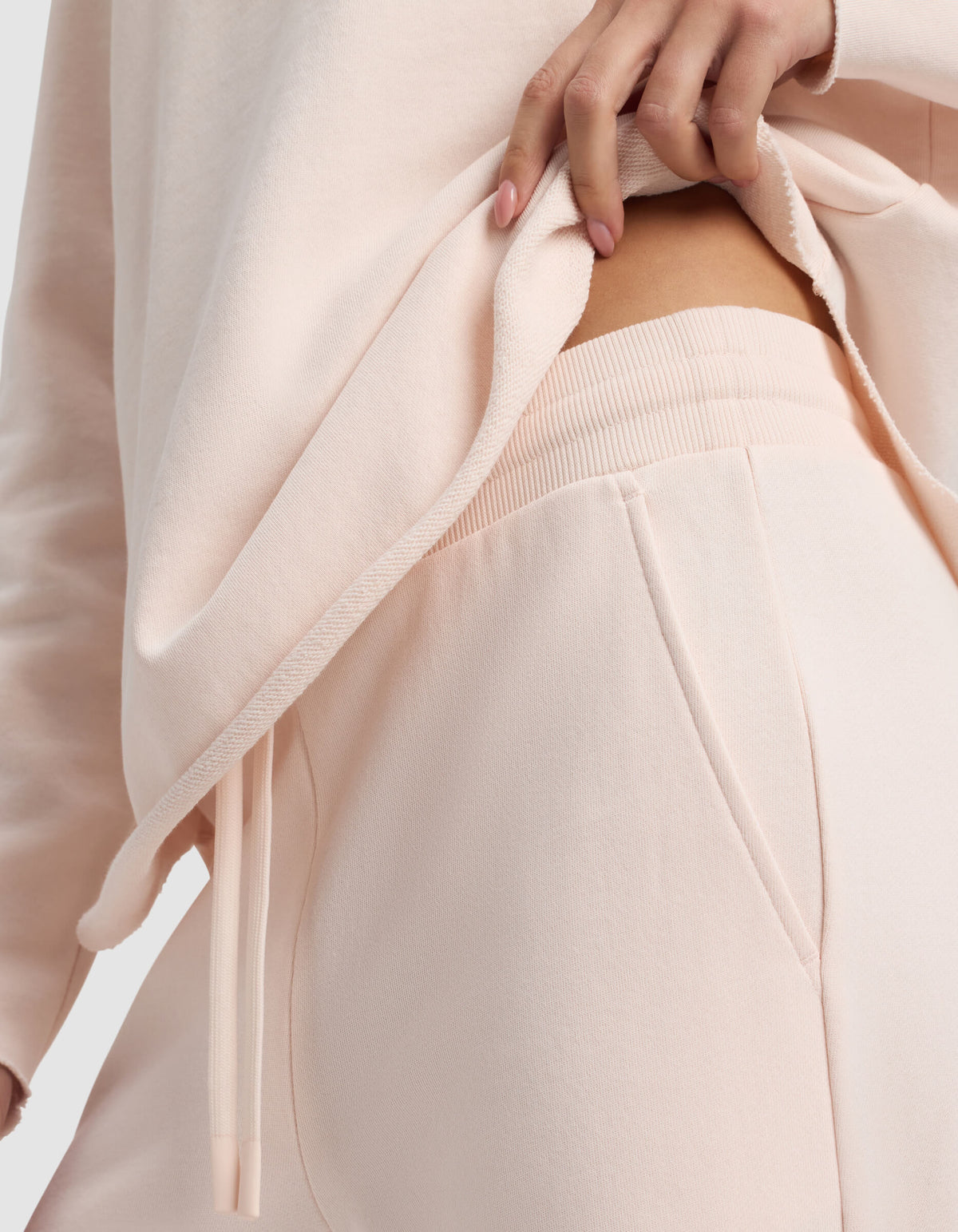A woman in Cozy Earth’s Women's Chelsea Cropped Pant lifts her matching top slightly, revealing the elastic waistband of the light pink pants.|Color:Petal