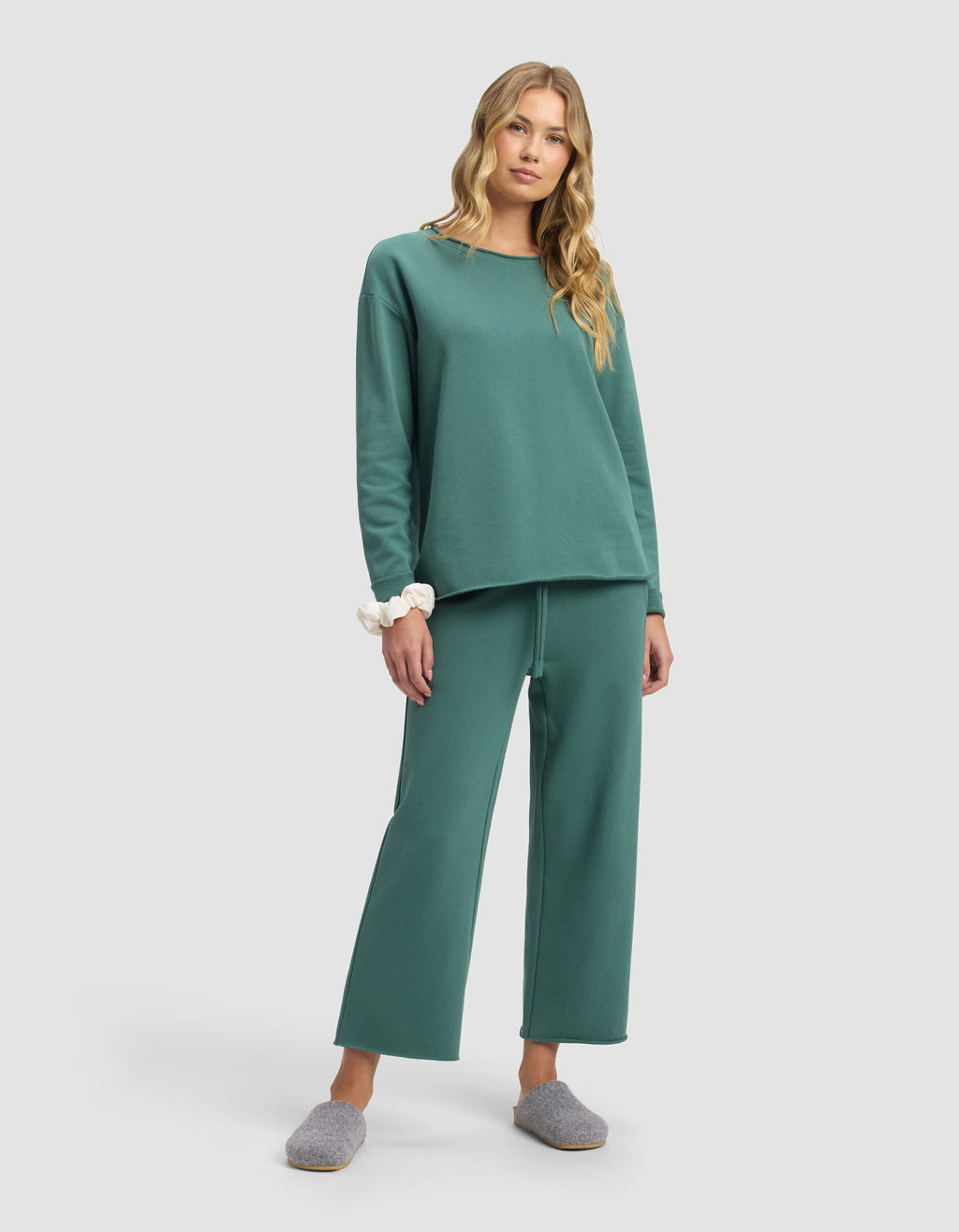 A woman with long blonde hair wears Cozy Earth’s Women’s Chelsea Cropped Pant in teal with a matching relaxed sweatshirt, a white scrunchie on her wrist, and gray slippers, standing against a plain light gray background.|Color:Sea Green