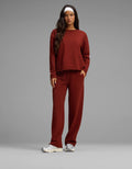 A woman stands against a plain gray background wearing the Cozy Earth Women's Chelsea Set—a matching rust-colored long-sleeve top and pants. She has long dark hair, a white headband, and white sneakers, with one hand in her pocket.|Color:Currant