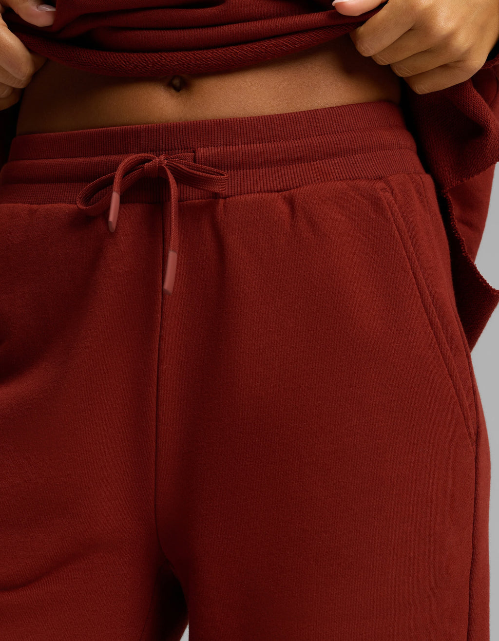 Close-up of a person wearing Cozy Earth’s Women's Chelsea Set, featuring high-waisted, drawstring maroon sweatpants and a matching maroon top, lifted to reveal part of their midriff and belly button.|Color:Currant