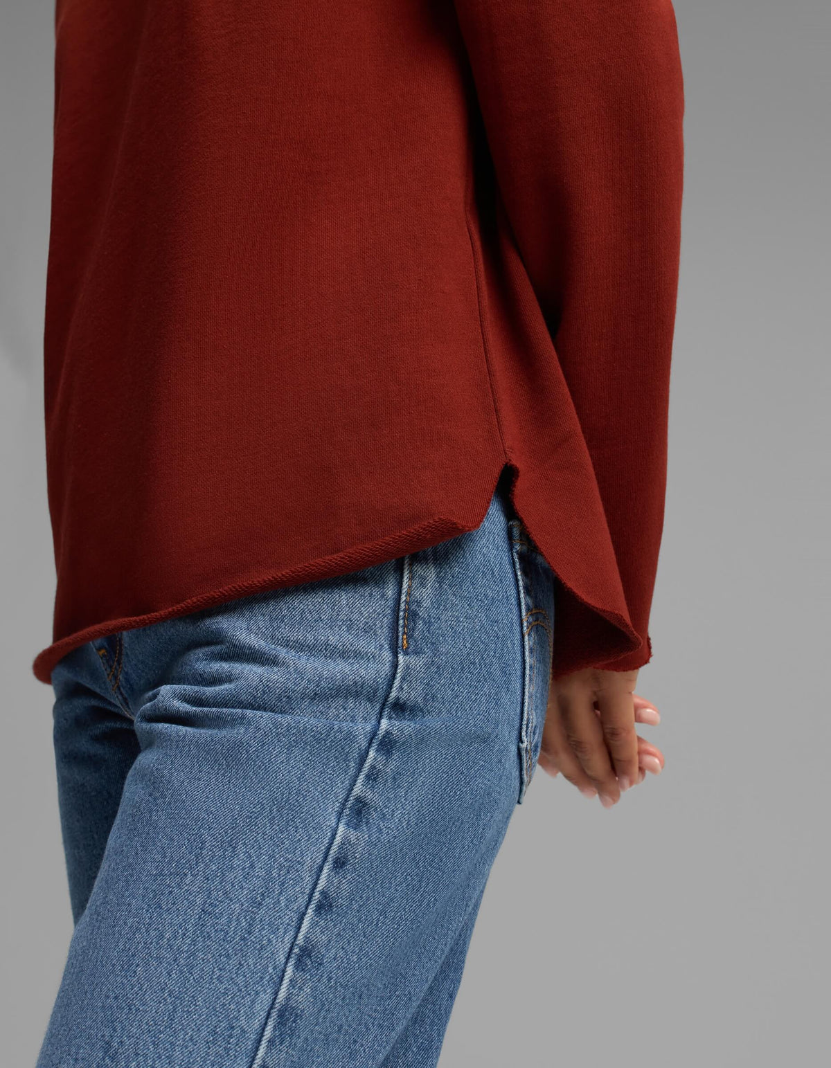 A person shown from the waist down, standing sideways with one hand behind their back against a plain gray background, wearing blue jeans and the Women's Chelsea Pullover in rust red by Cozy Earth.|Color:Currant
