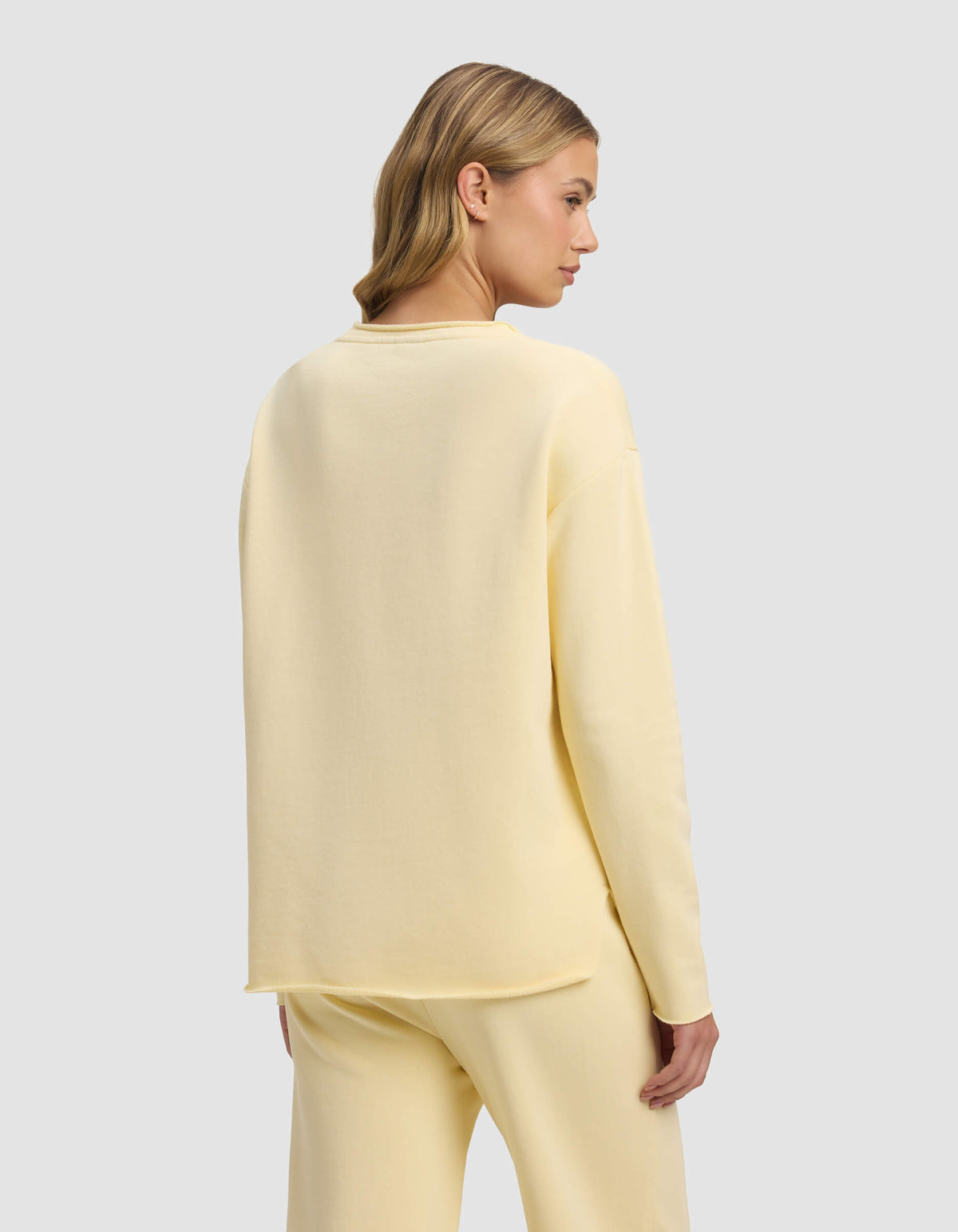 A woman with blonde hair styled in loose waves wears the Cozy Earth Women's Chelsea Pullover in pale yellow, paired with matching pants. She stands turned slightly away, displaying the back of her outfit against a light gray background.|Color:Meringue