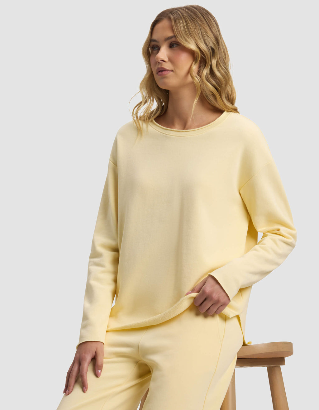 A woman with wavy blonde hair models the Cozy Earth Women's Chelsea Pullover in pastel yellow, paired with matching pants, as she sits on a wooden stool against a plain light gray background.|Color:Meringue