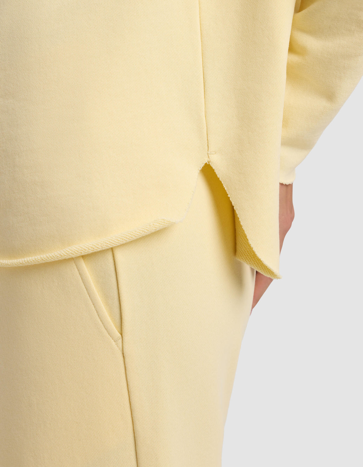 A close-up of a person wearing the Cozy Earth Women's Chelsea Pullover in pale yellow, featuring soft knit fabric, a curved hem with a side slit, and matching pants. Their hand is in the pocket against a plain light gray background.|Color:Meringue