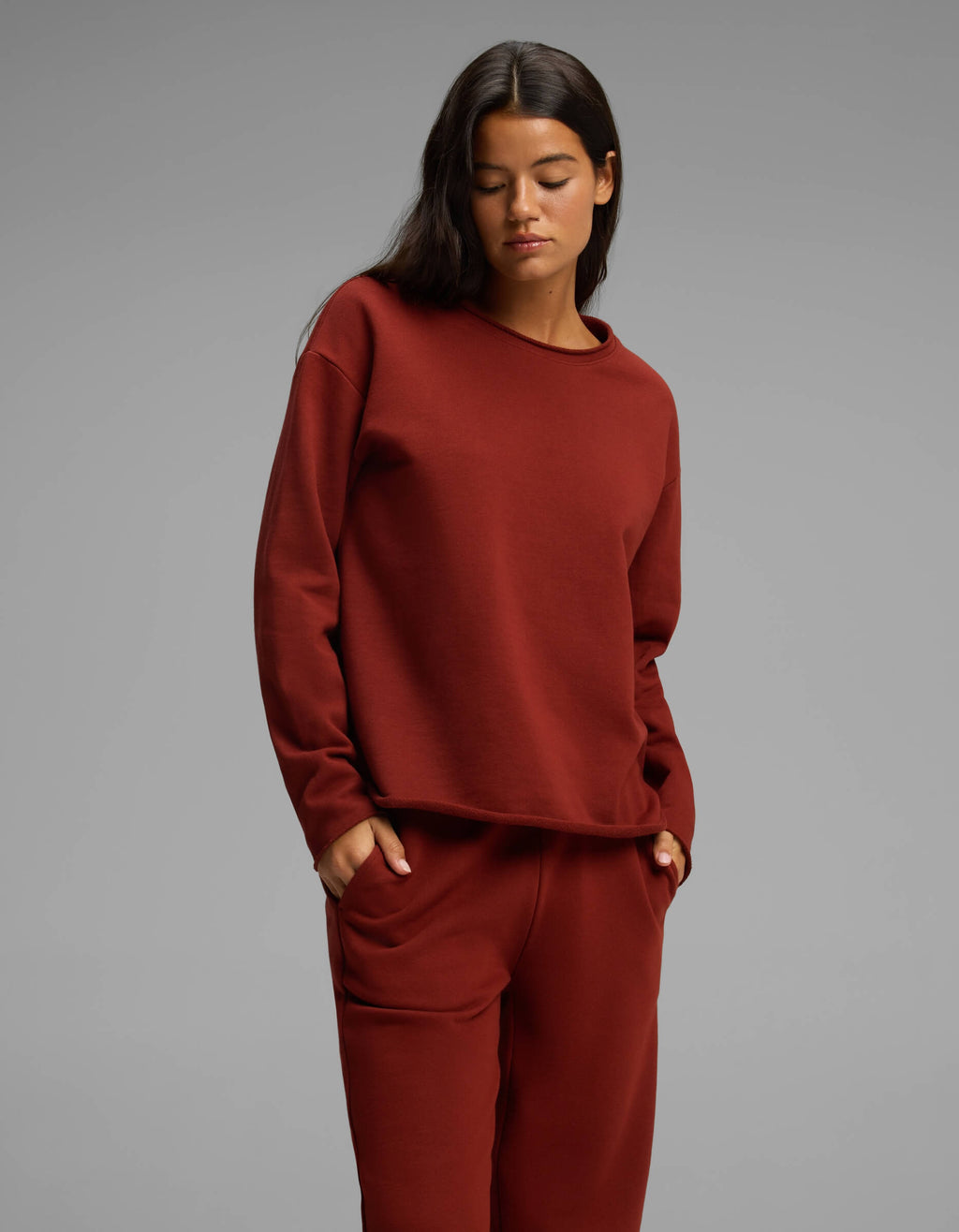 A person stands against a gray background wearing the matching maroon Women's Chelsea Set by Cozy Earth, with hands in pockets and looking down with a neutral expression.|Color:Currant