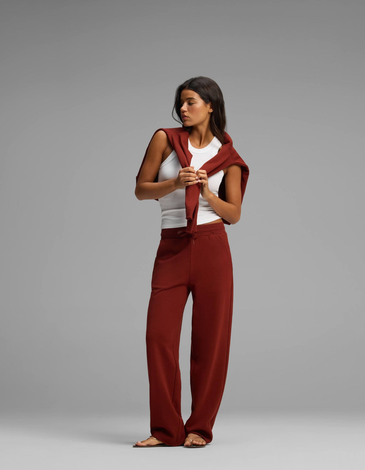 A woman stands against a gray background wearing the Women's Chelsea Set from Cozy Earth, which includes a white tank top, rust-colored pants, and a matching sweater draped over her shoulders. She looks down while holding the sweater’s sleeves in front of her.|Color:Currant