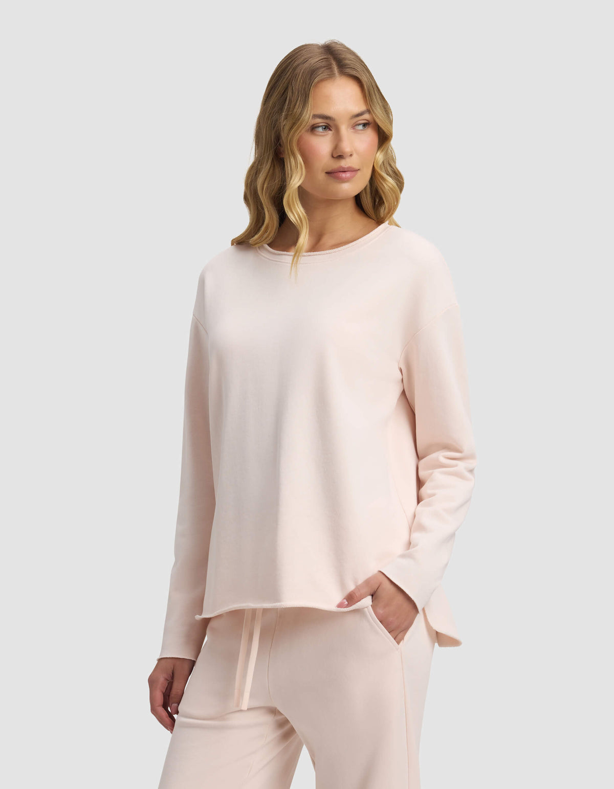 A woman with wavy blonde hair models the Cozy Earth Women's Chelsea Pullover in pale pink, paired with matching lounge pants, standing with one hand in her pocket against a plain light gray background.|Color:Petal