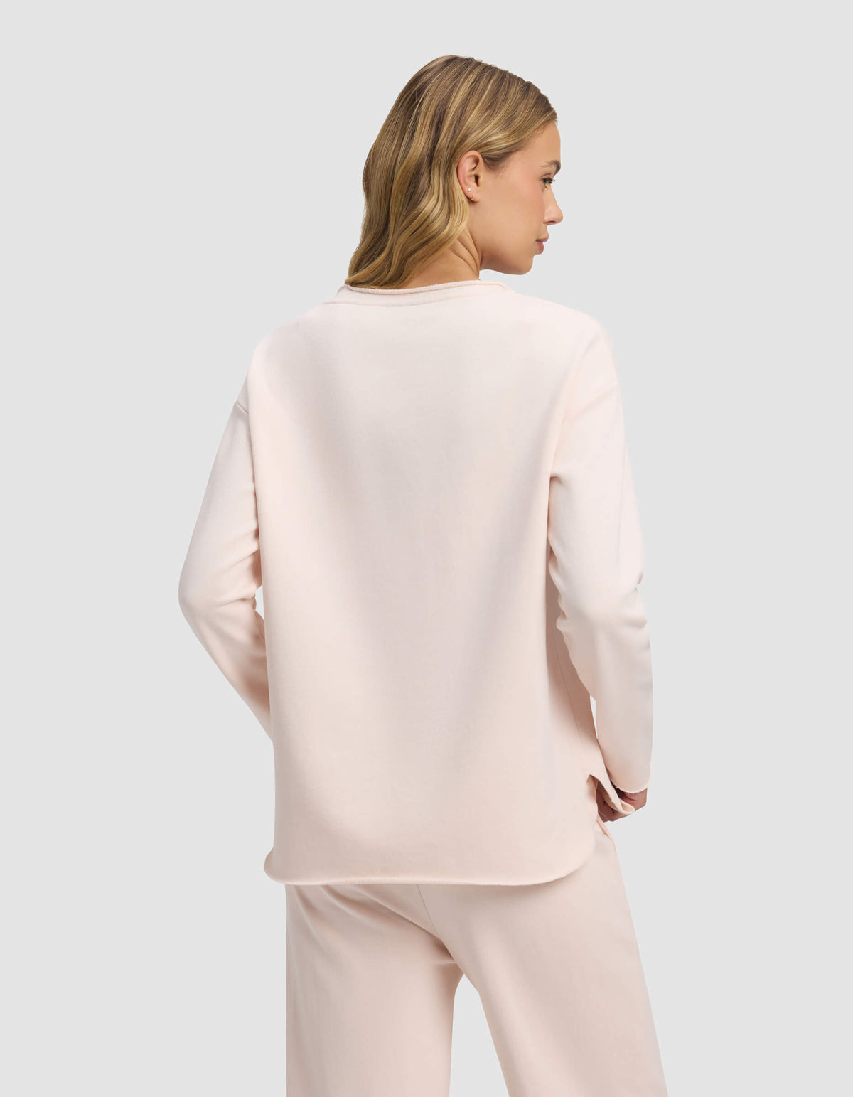 A woman with blonde hair models the Cozy Earth Women's Chelsea Pullover in light pink, standing with her back to the camera against a plain light grey background.|Color:Petal